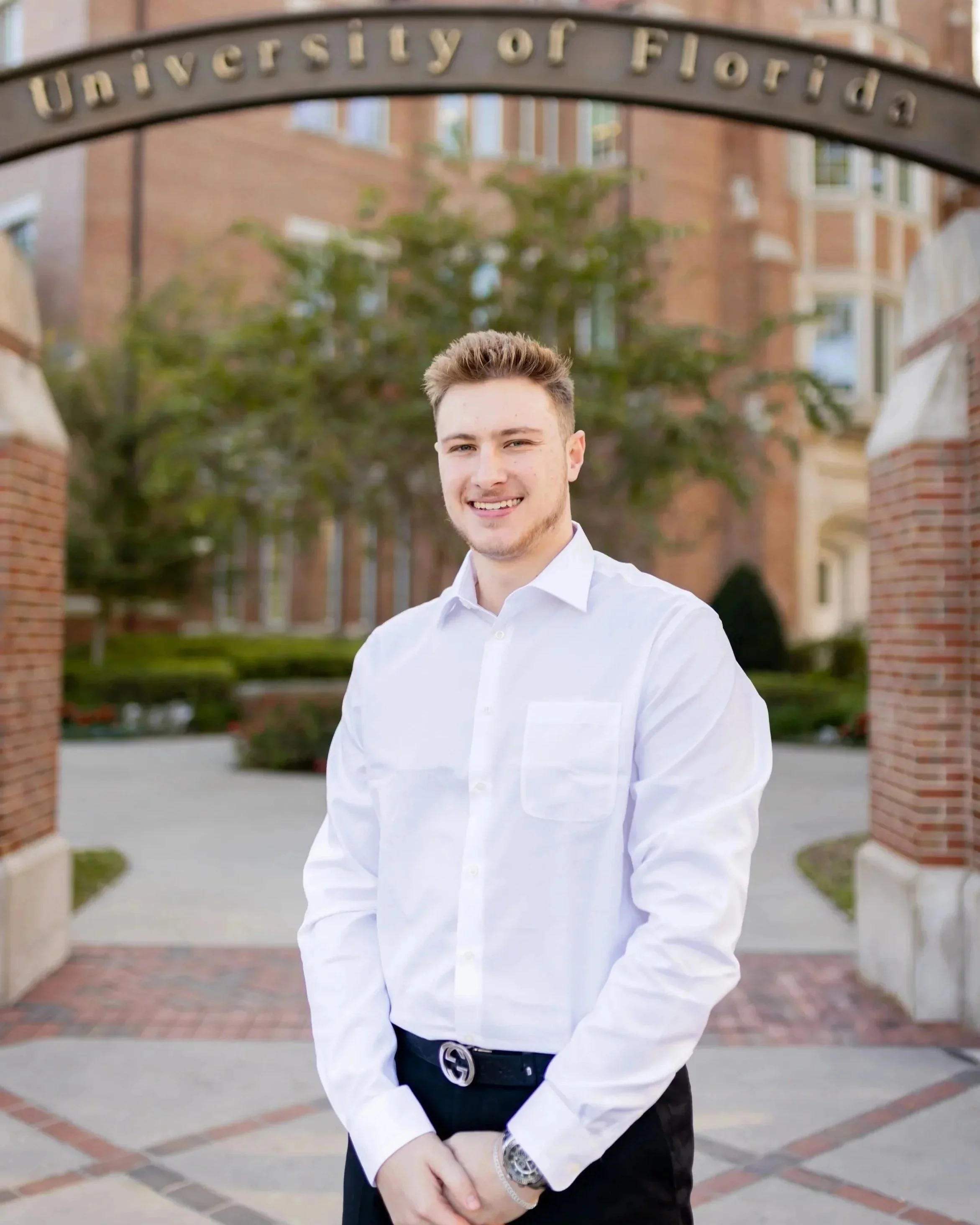 jonathan odom with short light-colored hair smiling, wearing a white dress shirt and black belt, standing outdoors in front of a university building with an arch that reads 'University of Florida' and green trees in the background.