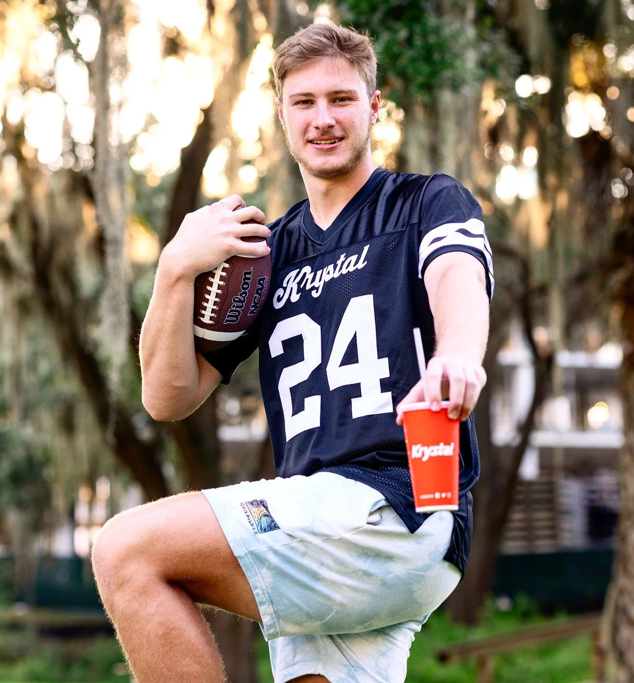 A young man outdoors holding a football and a red cup. He is smiling, wearing a black football jersey with the number 24 and the logo 'Krystal,' and white shorts.