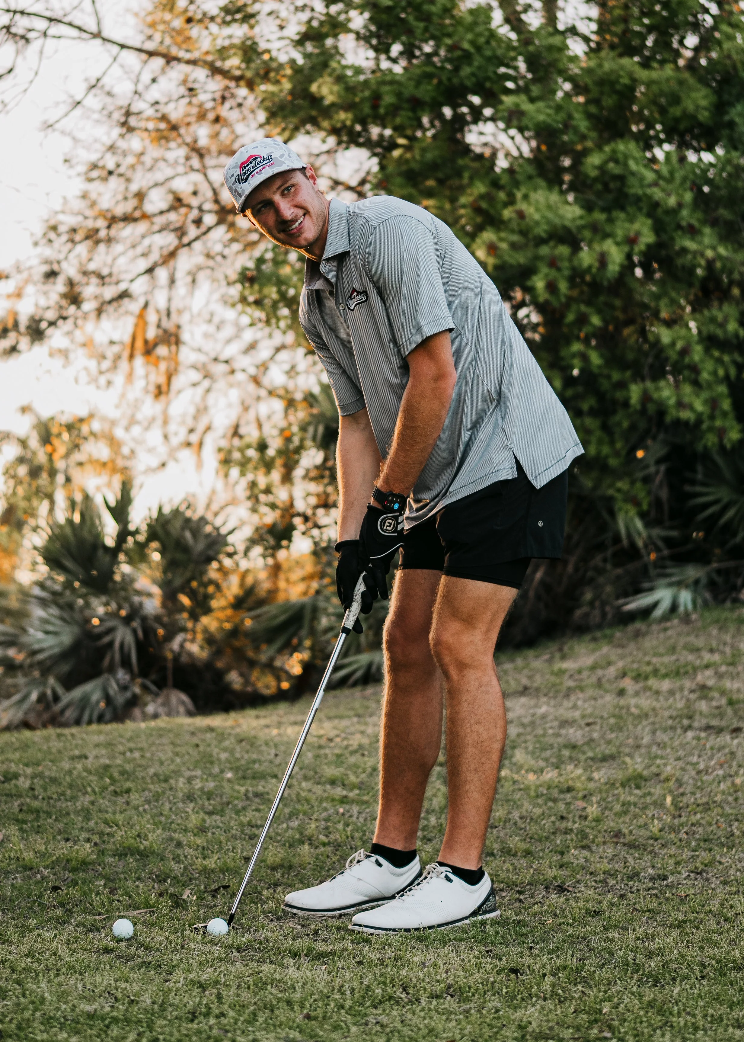 Jonathan Odom playing golf outdoors, wearing a hat, gray shirt, black shorts, and white golf shoes, preparing to hit a golf ball with a club, with trees and bushes in the background during sunset.