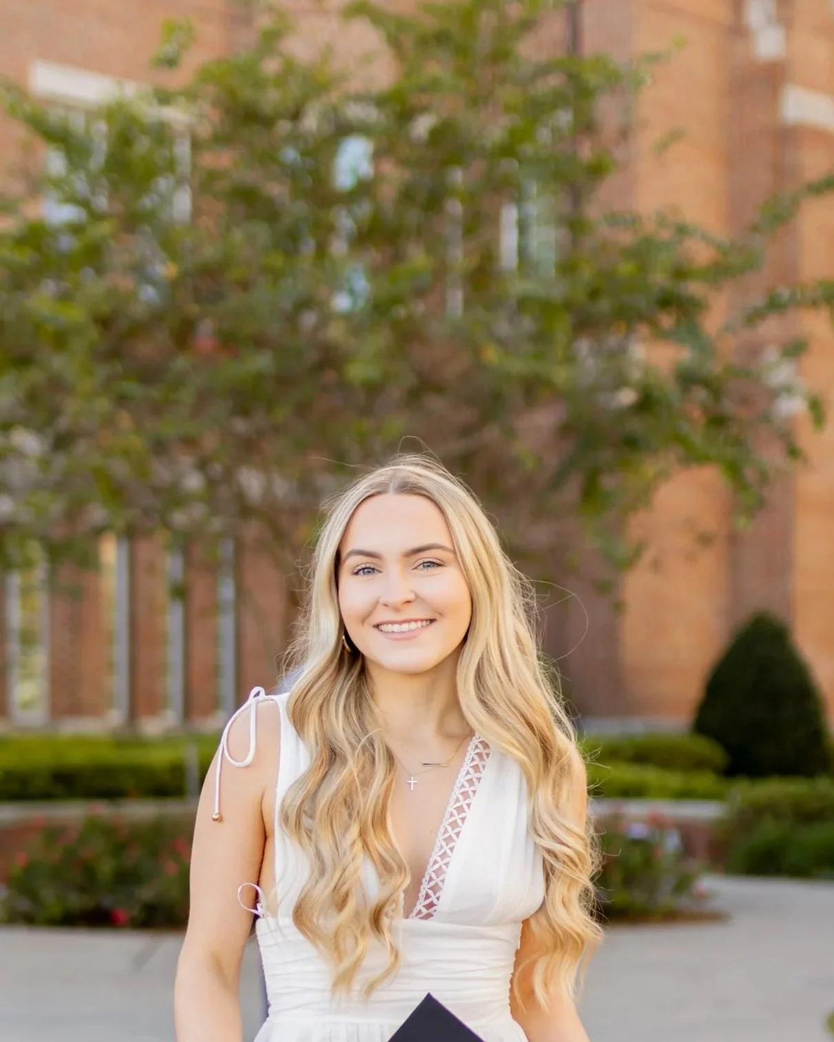 A young woman with long blonde hair, smiling, wearing a white dress, standing outdoors with a brick building and trees in the background.