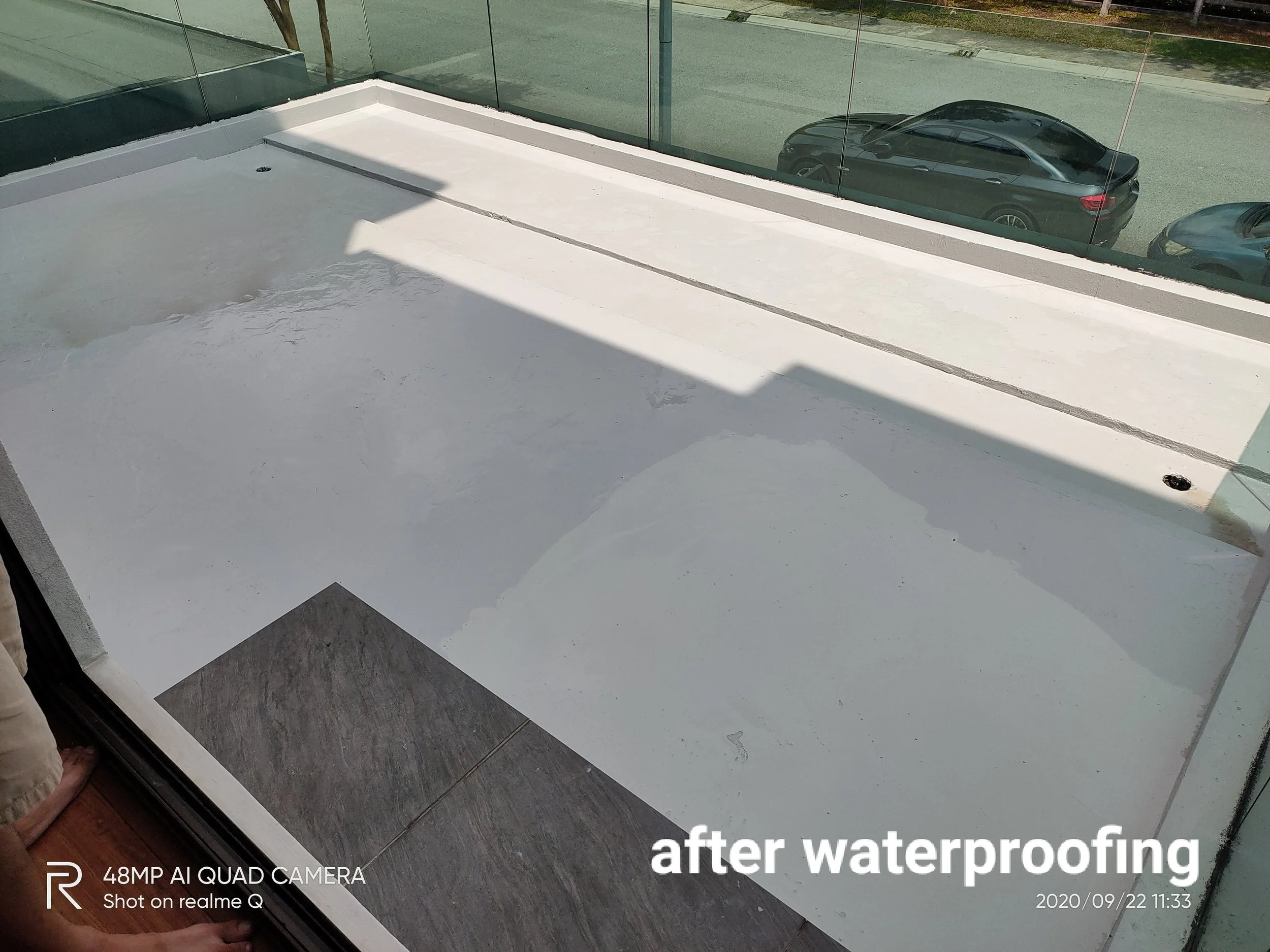 Photo showing a balcony with water-resistant coating, surrounding glass panels, and parked cars visible outside. The text on the image indicates the waterproof coating was done after waterproofing.