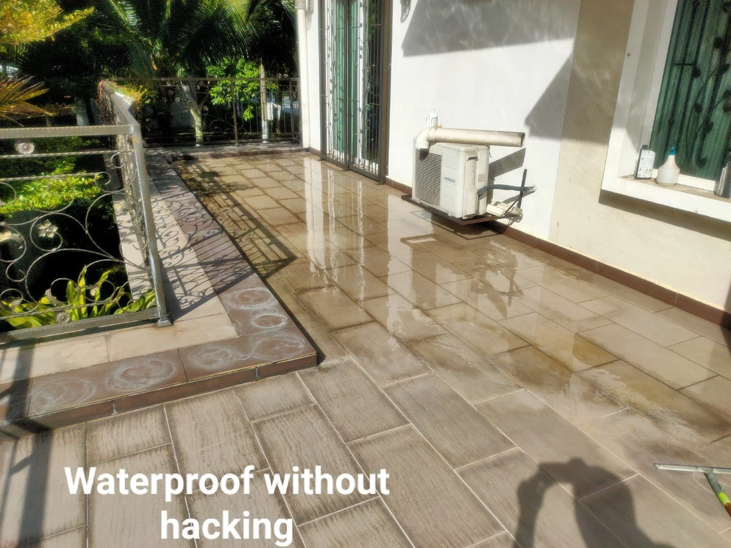 A patio or balcony with a wet tiled floor, a metal railing on the left, a white wall with windows on the right, and an air conditioning unit. The image has a shadow of a person taking the photo, and the caption says 'Waterproof without hacking'.