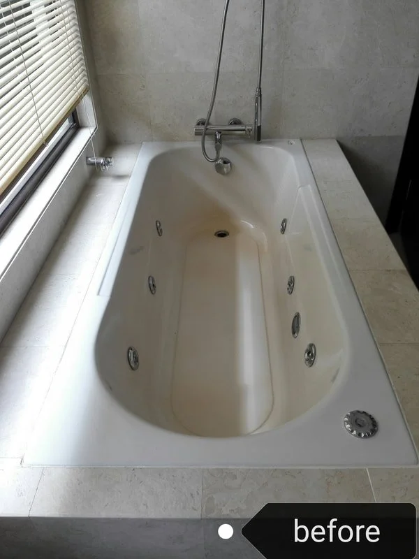 A white bathtub with multiple jet nozzles, positioned next to a window with blinds, in a bathroom with beige tiled walls and floor, ready for a makeover.