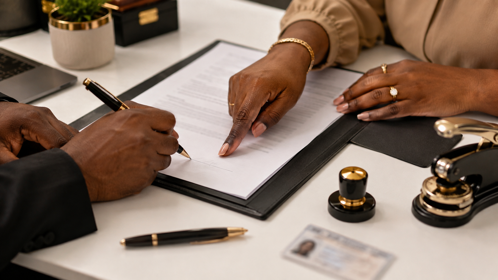 Two people signing a document at a desk, with a woman pointing at the paper, various office supplies, a plant, and a photo ID visible on the desk.