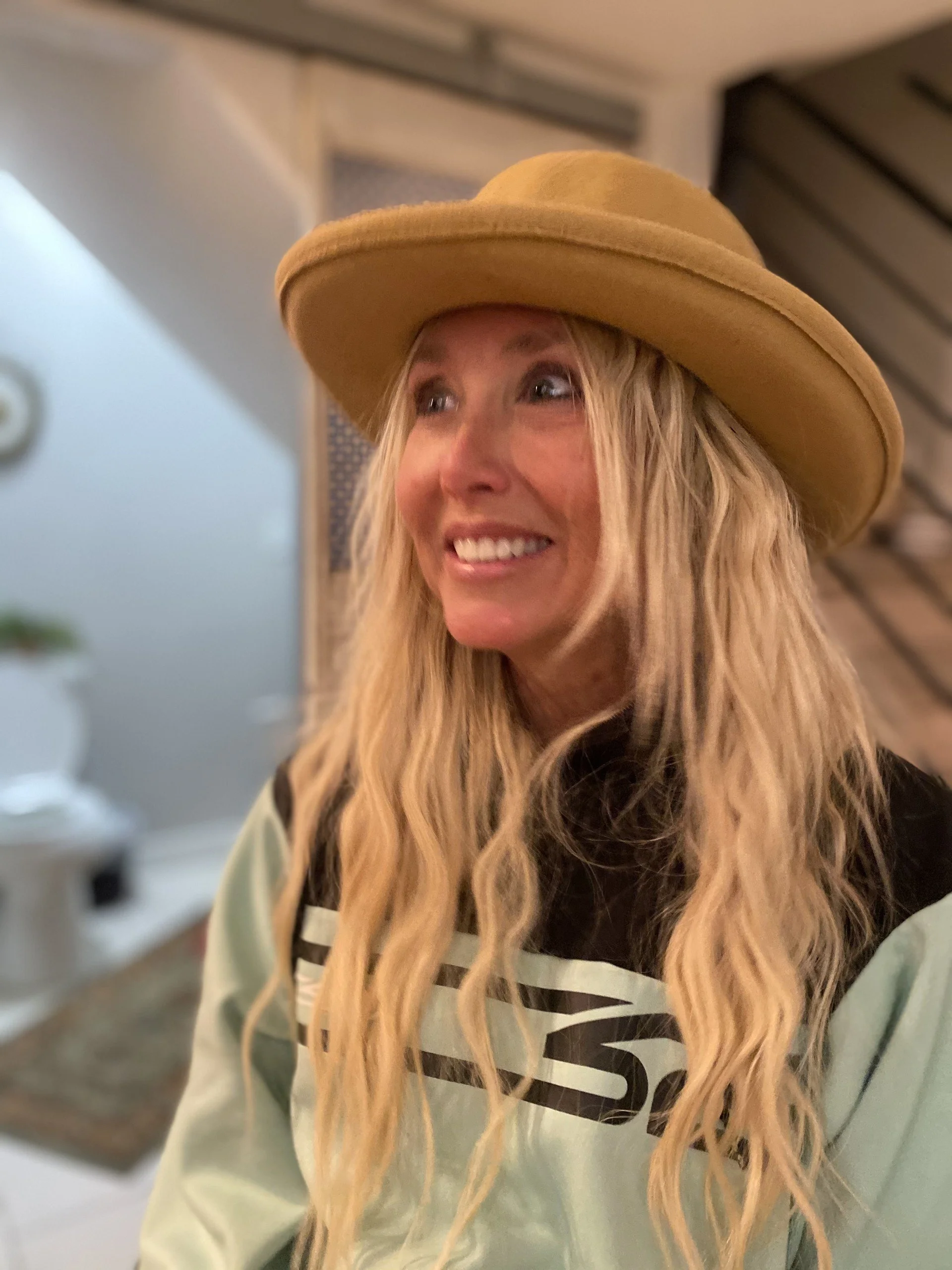 A woman with long, wavy blonde hair wearing a wide-brimmed tan hat and a striped shirt, smiling and looking to the side indoors.