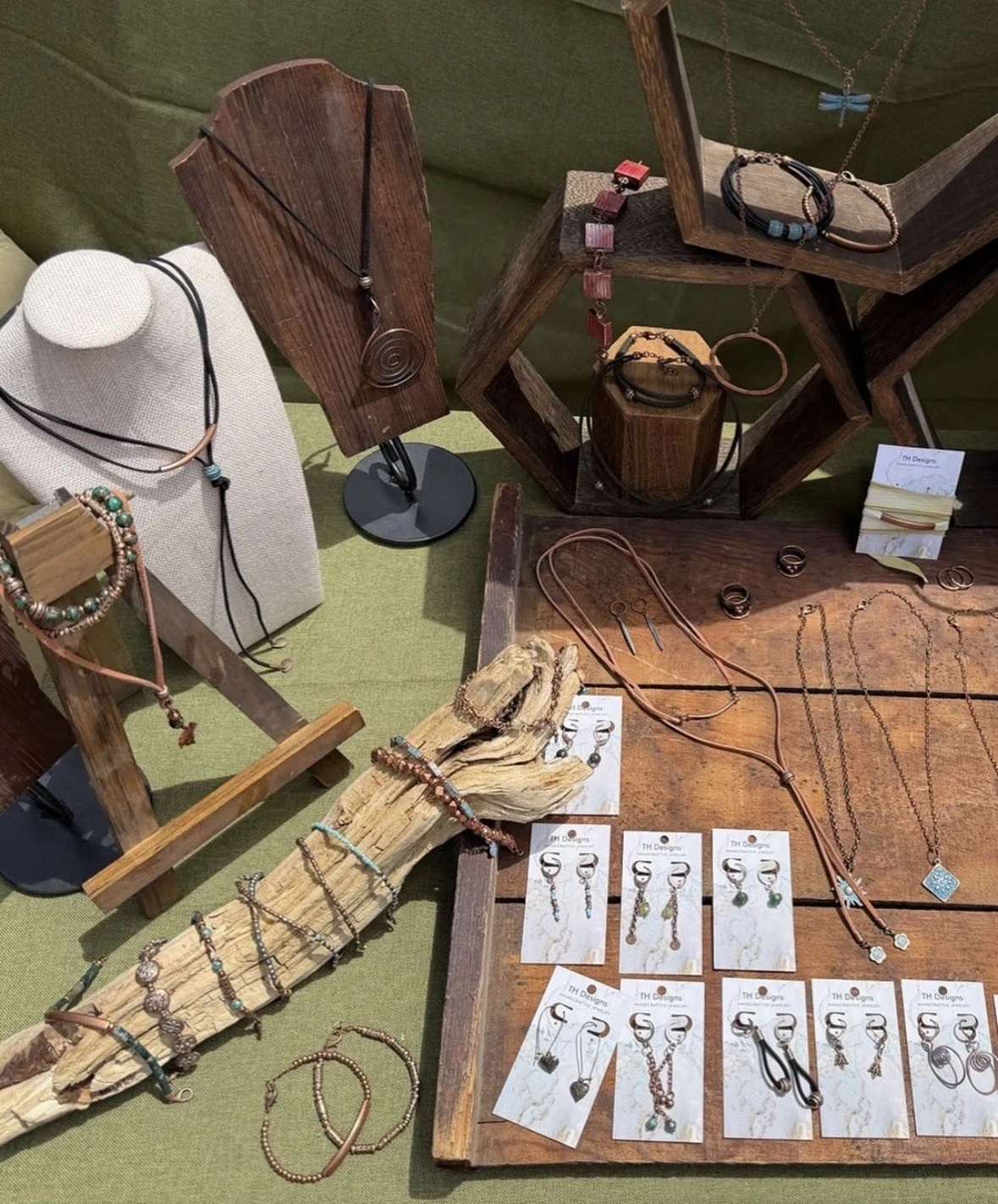 Display of various handmade jewelry pieces including necklaces, bracelets, earrings, and rings, arranged on wooden and fabric surfaces at a craft fair or boutique shop.