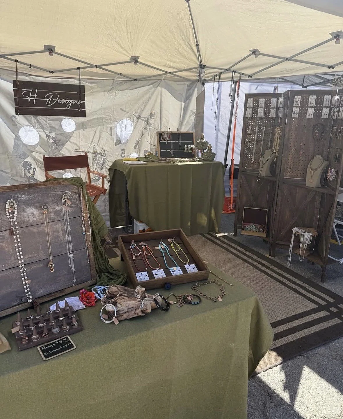 Jewelry display booth with necklaces and bracelets, wooden jewelry organizers, and a sign that says 'H Designers' inside a tent.