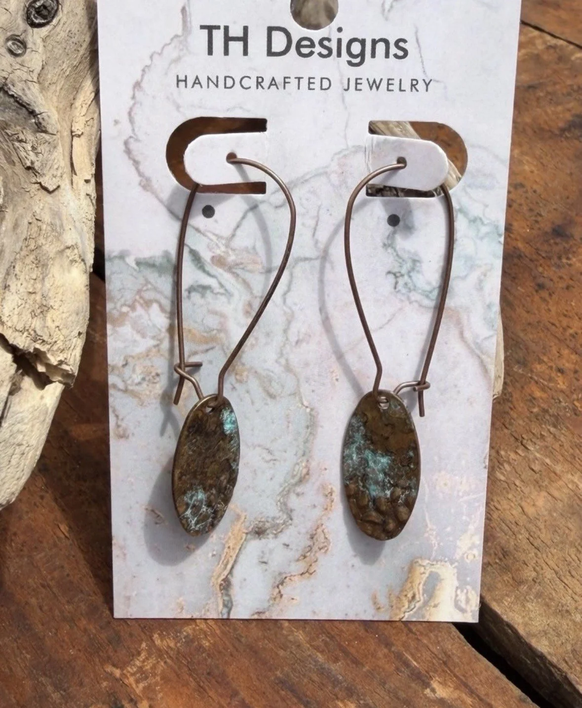A pair of handcrafted jewelry earrings with elongated metal wire hooks and oval-shaped stones with a rustic, earthy appearance, displayed on a card from TH Designs.