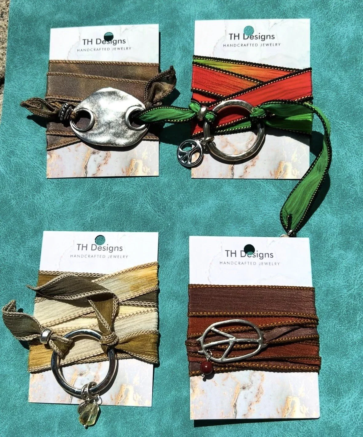 Four handmade fabric bracelets with metal accents on display on a green surface. Each bracelet is attached to a white card from TH Designs, featuring a variety of colors and designs.