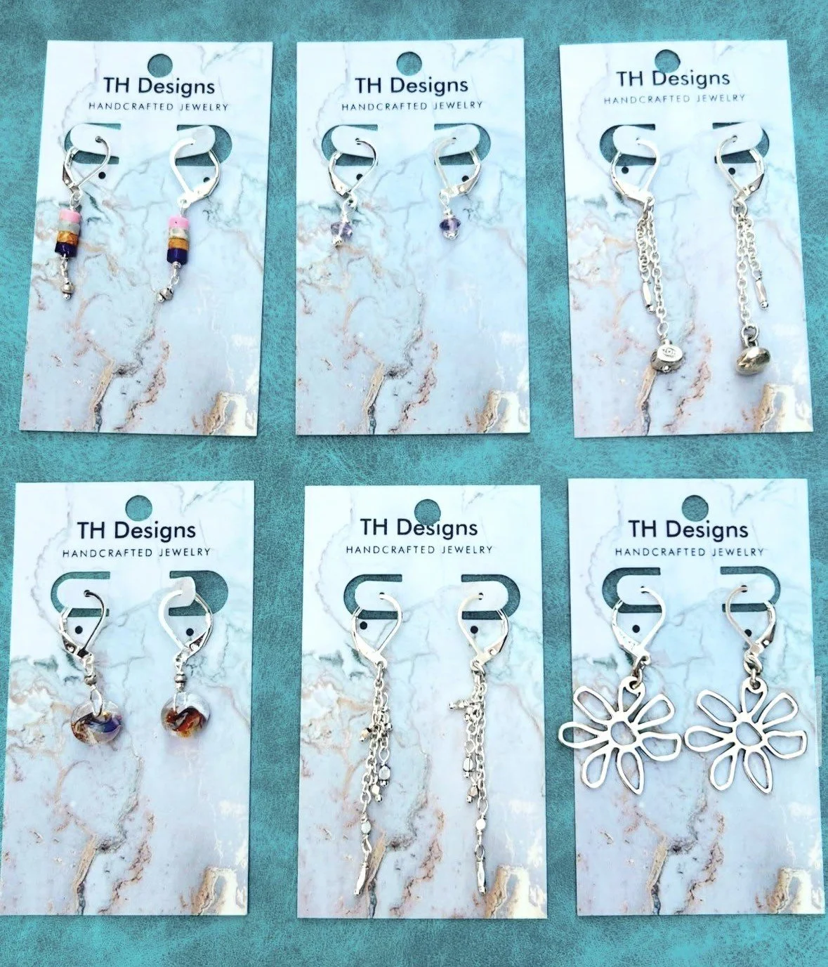 Six pairs of handcrafted earrings on white cards labeled 'TH Designs Handcrafted Jewelry' arranged on a teal surface. The earrings vary in design, including beads, chains, and floral shapes.