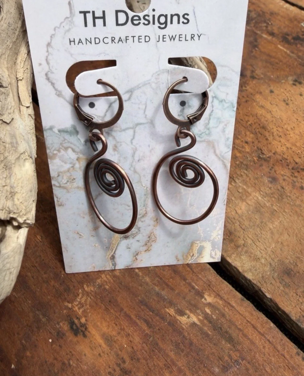 A pair of handmade wire earrings with spiral designs, attached to a card labeled 'TH Designs' that advertises handcrafted jewelry, placed on a wooden surface.