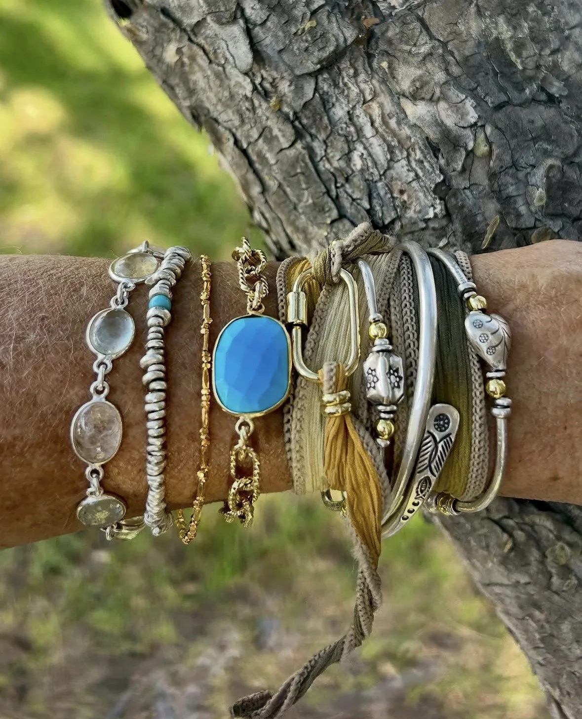 A person's wrist adorned with multiple jewelry bracelets in silver, gold, and mixed metals, wearing various designs including charms, beads, and a large blue gemstone, against a background of a tree trunk and green foliage.