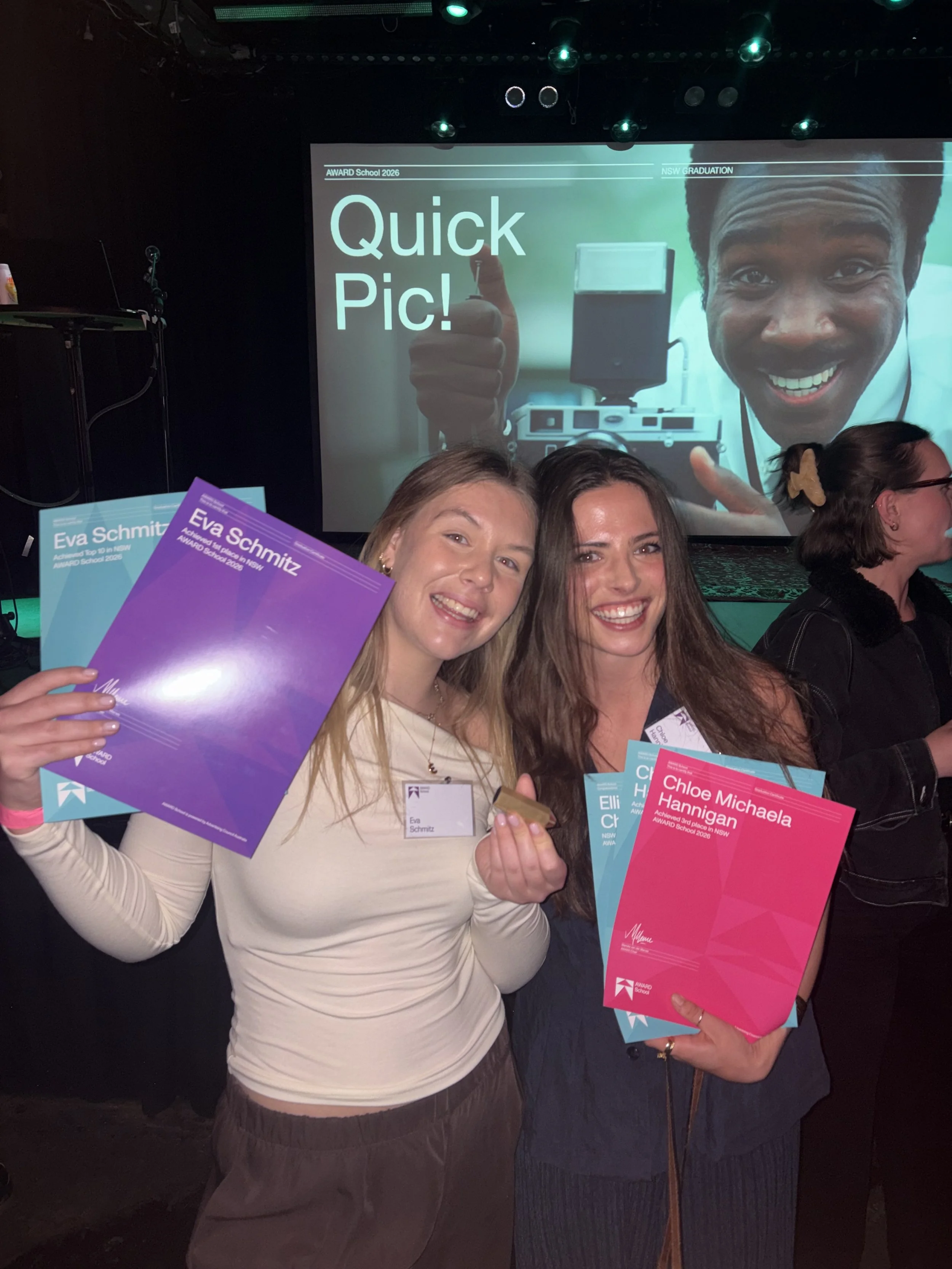 Two young women smiling and holding colorful award certificates at a graduation event, with a stage and a large screen displaying a man giving a thumbs-up in the background.
