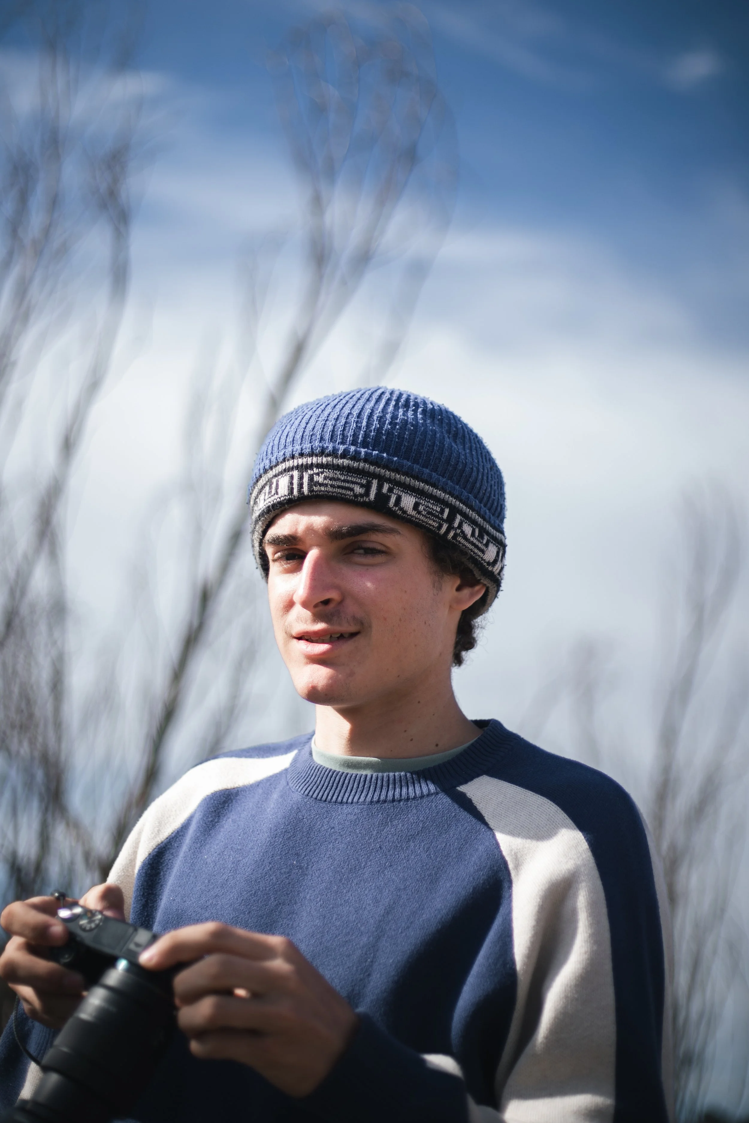 Young man outdoors wearing blue and gray sweater and a blue knit hat, holding a camera in his hands, with bare trees and a cloudy sky in the background.