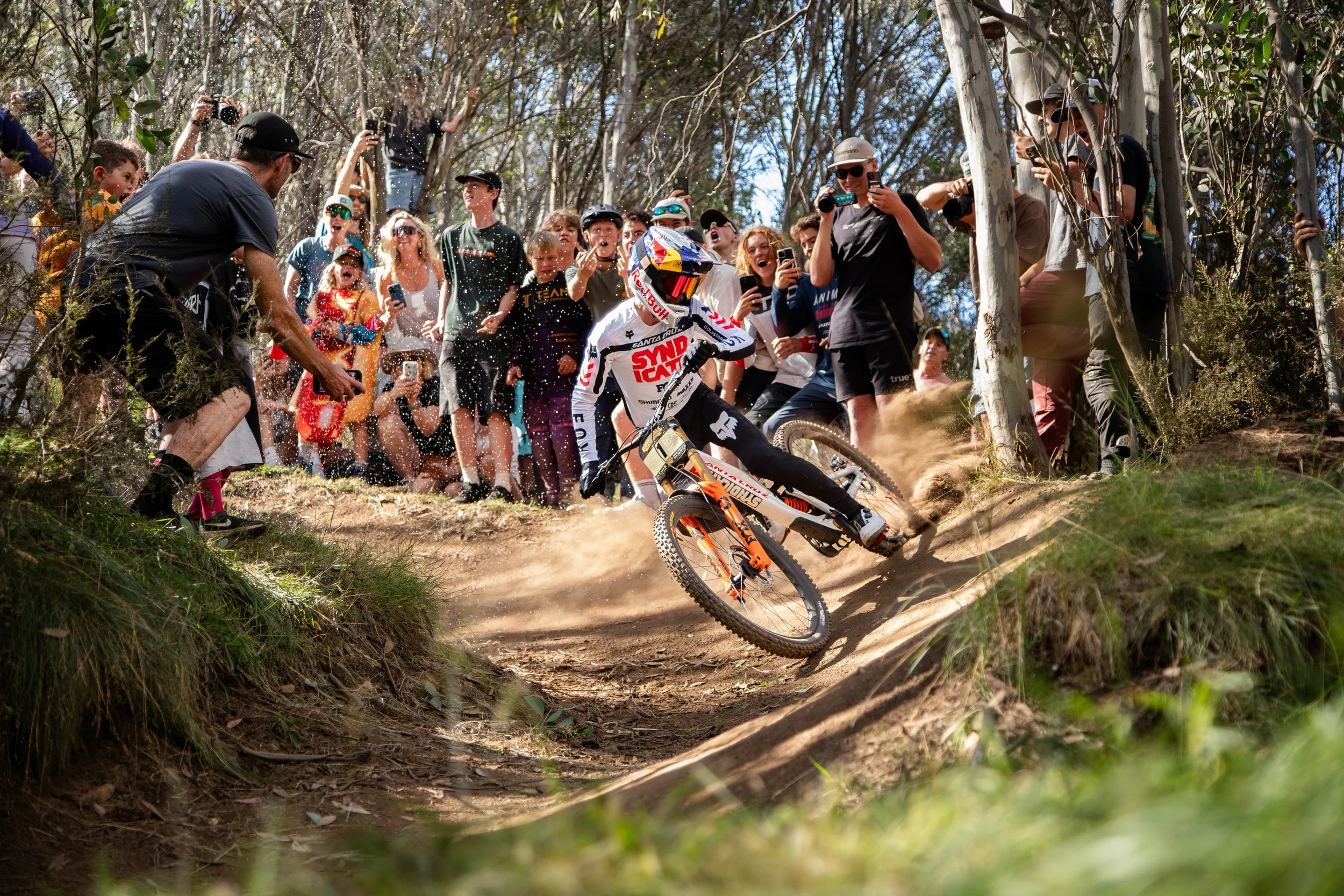 A mountain biker in white racing gear with a full-face helmet riding downhill on a dirt trail in a forest, surrounded by a crowd of spectators capturing the moment with their phones and cameras. The spectators are standing behind trees and on the hillside, many smiling and cheering.
