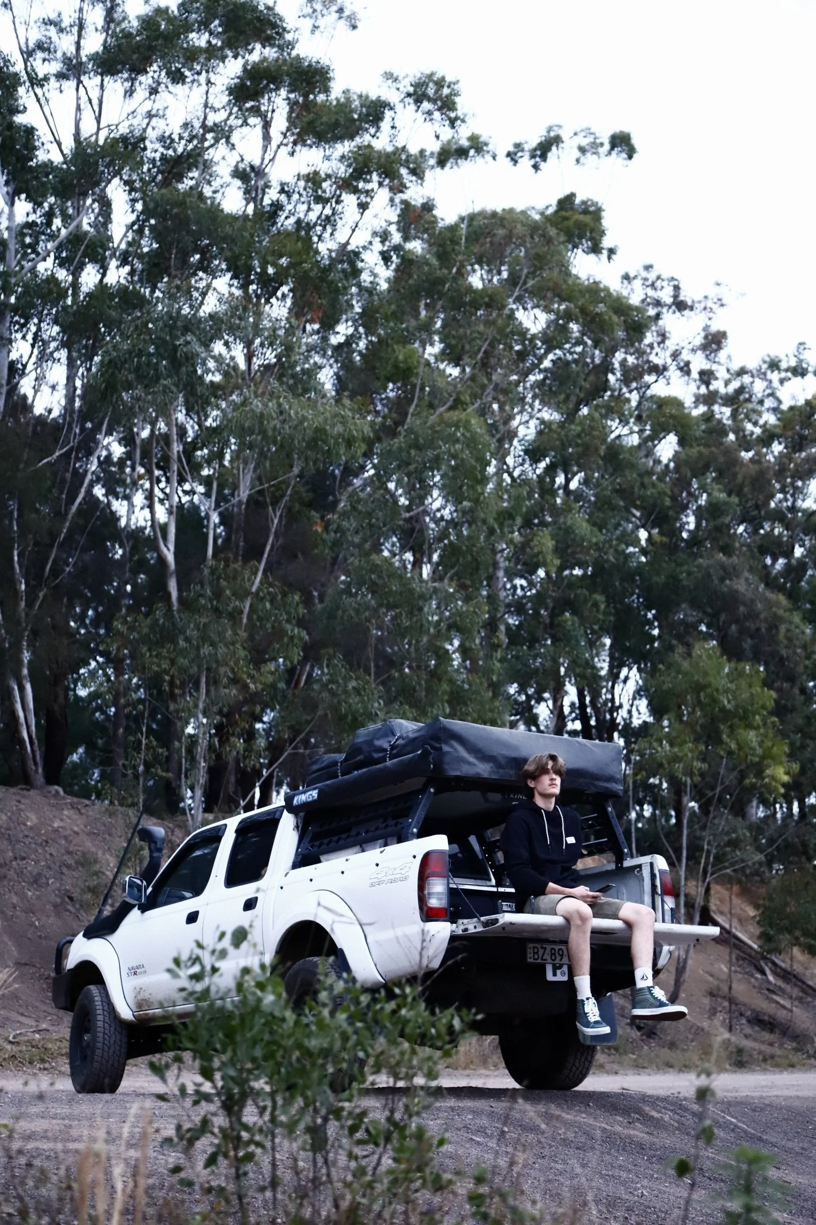 A young man sitting on the tailgate of a white pickup truck with a rooftop tent, parked on a dirt road in a wooded area with tall trees and a clear sky.