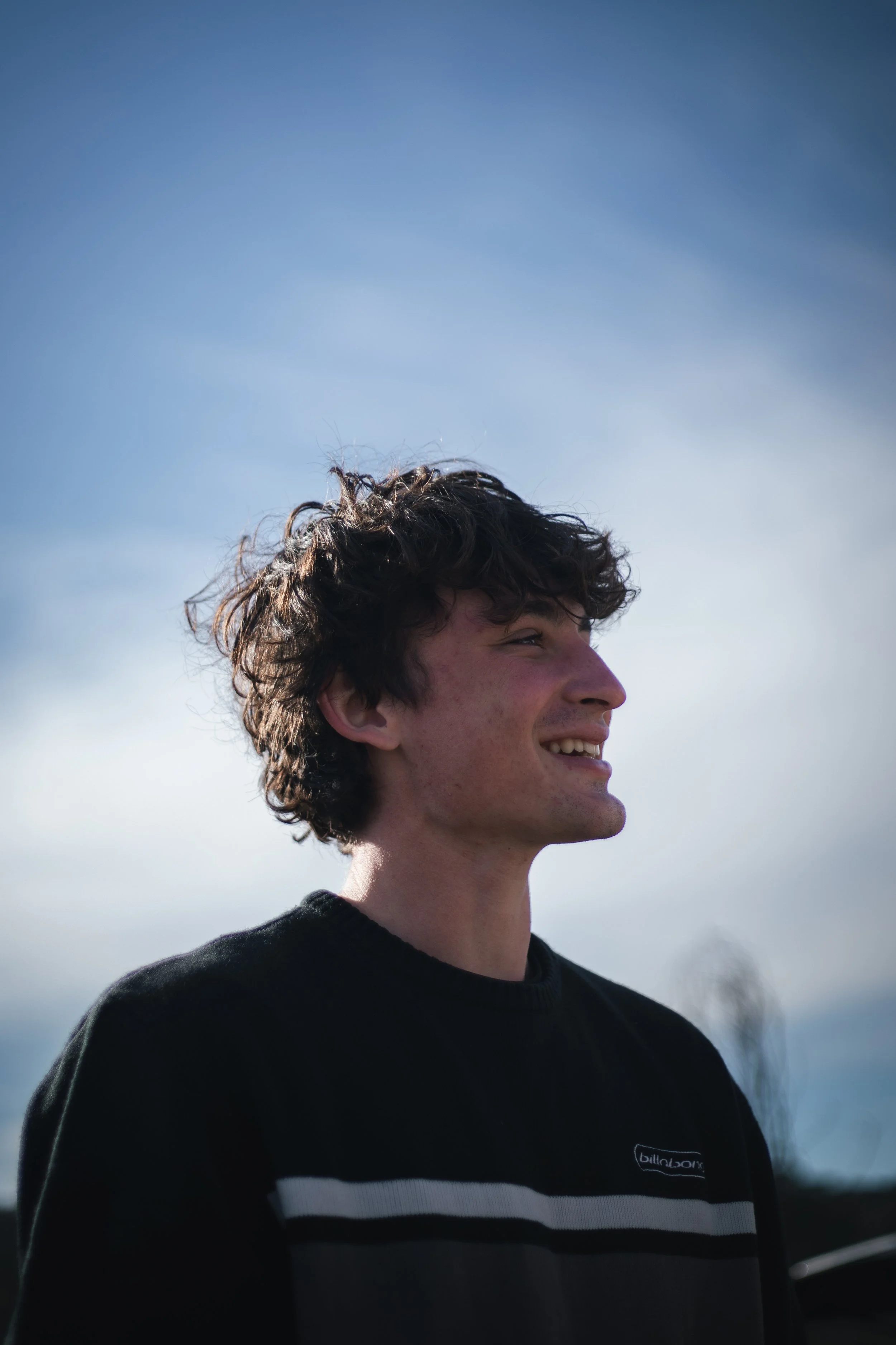 profile of a young man smiling outdoors with a cloudy sky in the background, wearing a black sweater with white horizontal stripes.