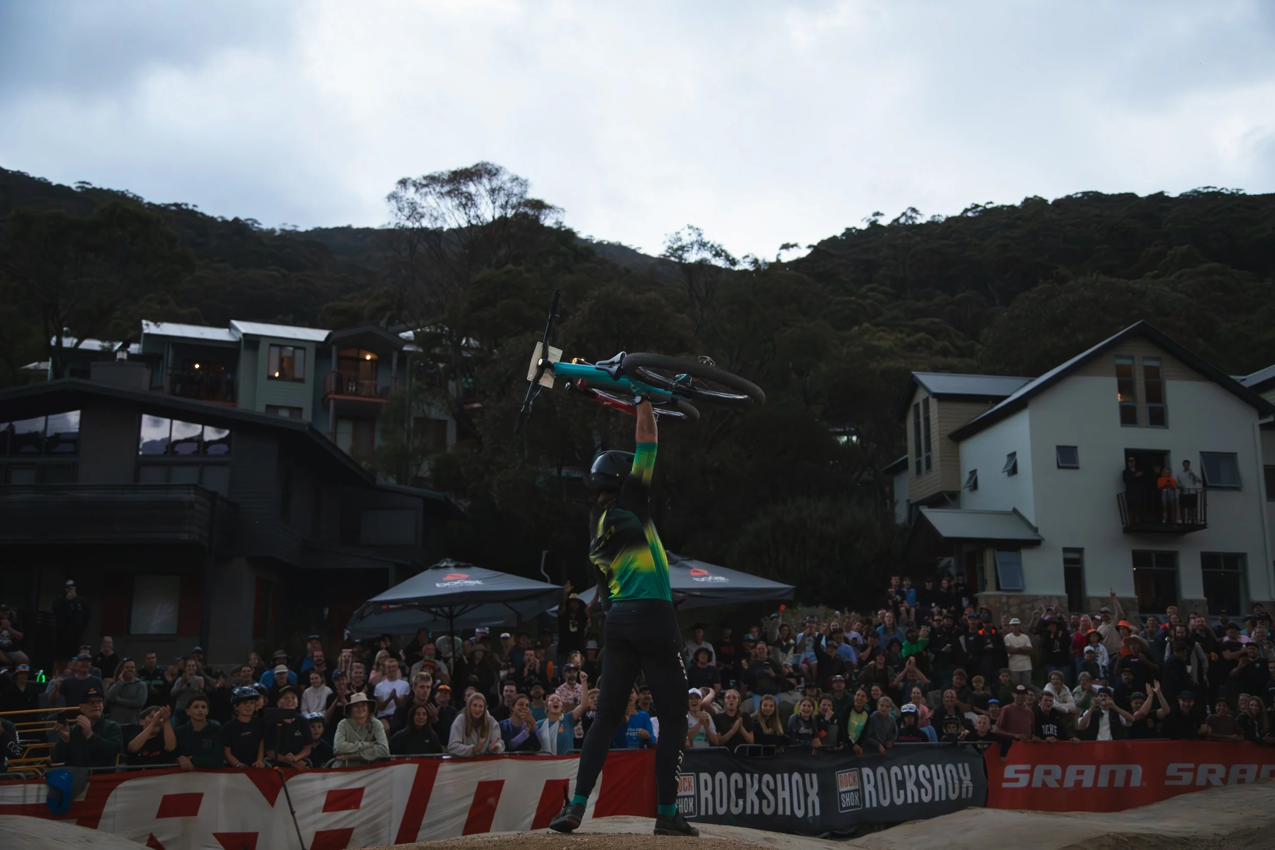 A cyclist performing a stunt involves a headstand on a bike, with an audience watching at an outdoor event with a backdrop of houses, trees, and a cloudy sky.
