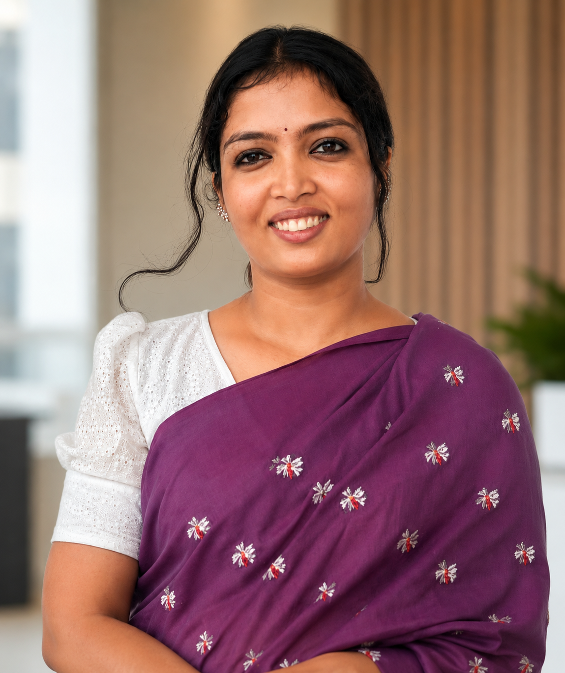 A smiling woman with dark hair, wearing a white blouse and a purple sari with small embroidered designs, standing indoors with blurred background.