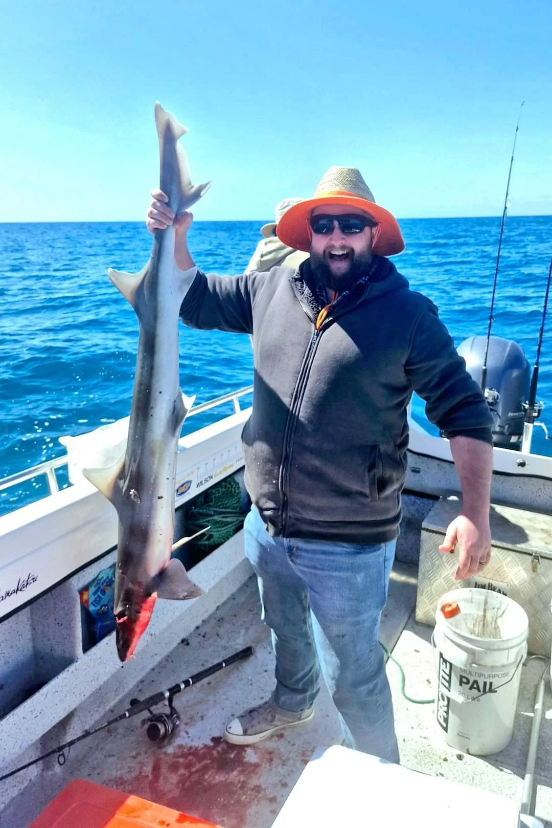 A man on a boat holding a large fish, smiling, wearing sunglasses, a wide-brimmed hat, a hoodie, and jeans, with the ocean and blue sky in the background.