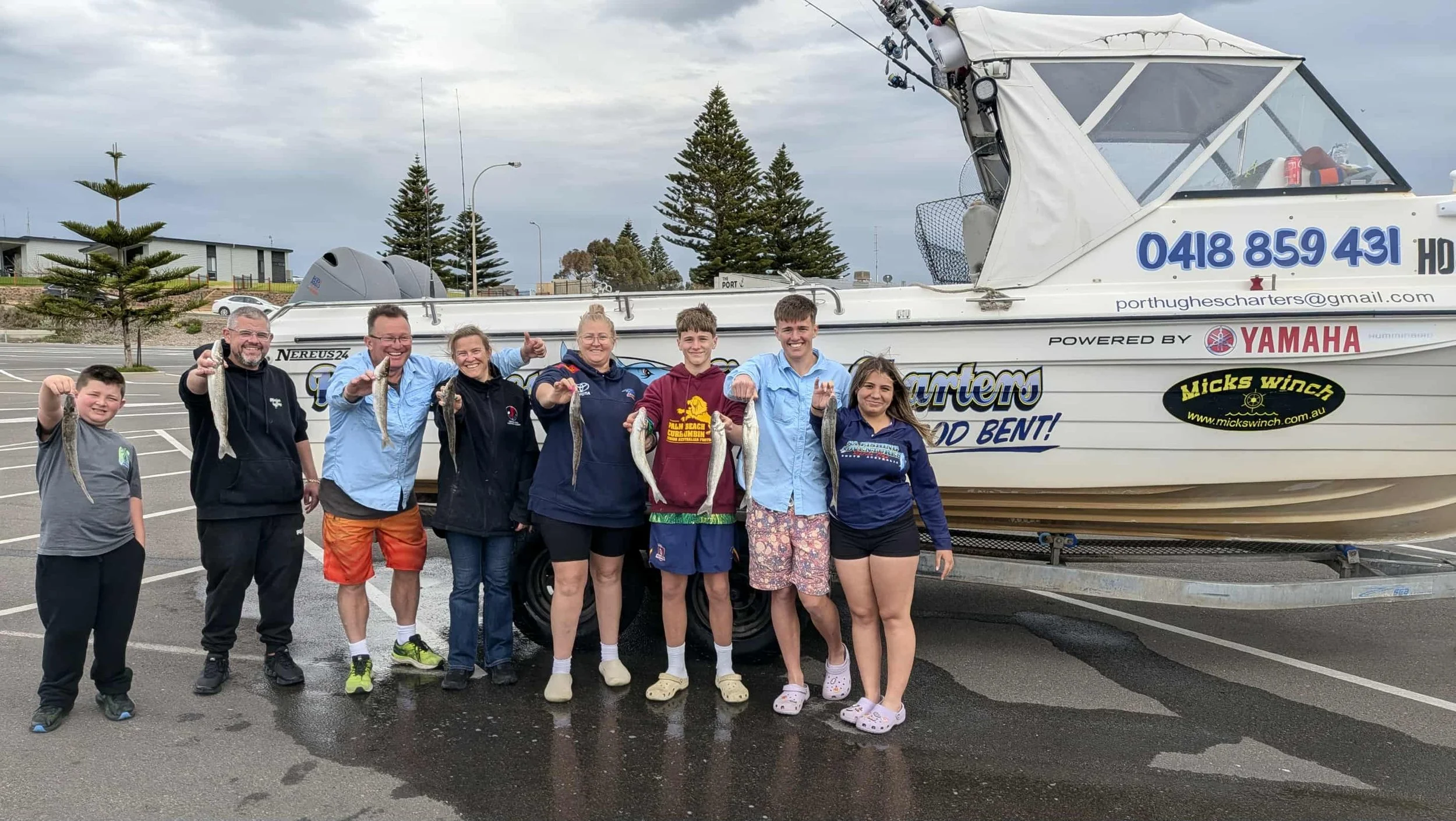 A group of nine people standing in a parking lot holding fish in front of a white boat on a trailer, with trees and a cloudy sky in the background.