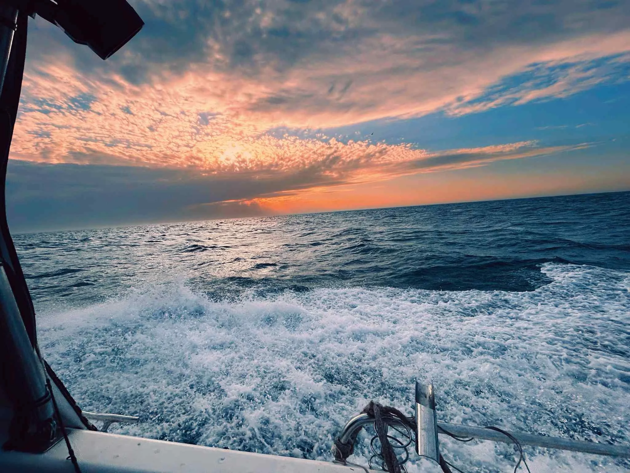 View of the ocean at sunset, with a colorful sky filled with pink, orange, and blue clouds. The photo appears to be taken from a boat moving through the water, leaving a wake behind.