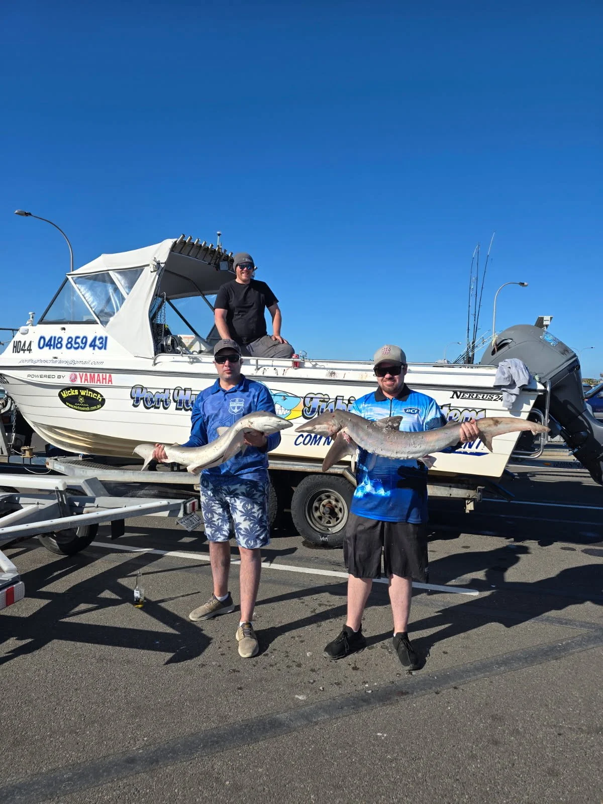 Two men holding large fish in front of a sailboat on a trailer, with a third man sitting on the boat, during a sunny day.