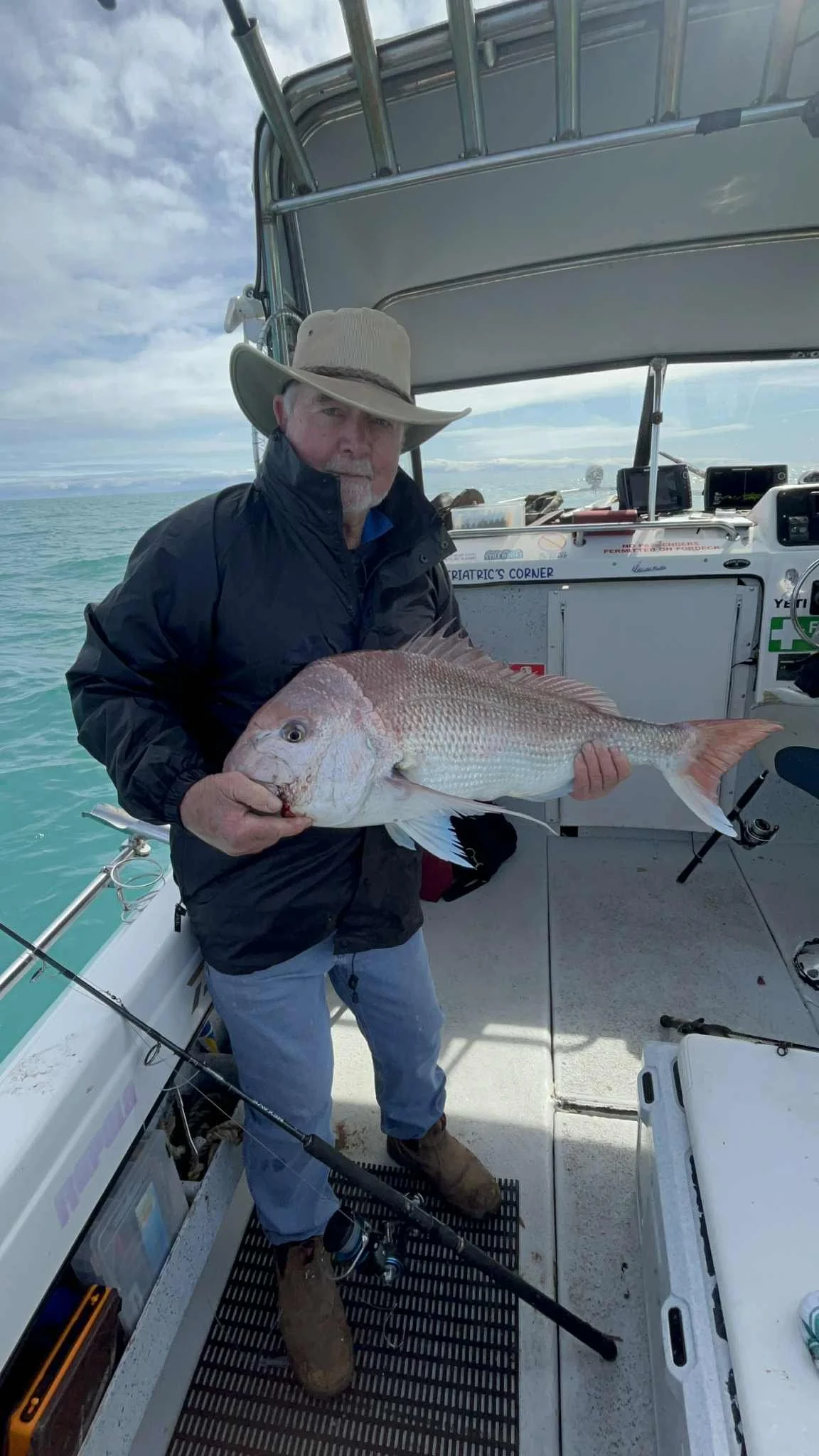 A man wearing a wide-brimmed hat, black jacket, jeans, and boots holding a large fish aboard a boat on the water.
