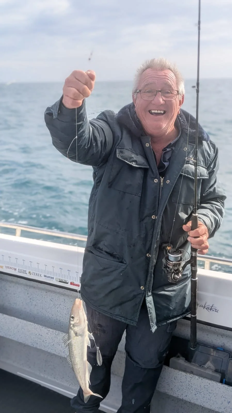 An older man with glasses, smiling widely, holds a fishing rod with a fish he caught, on a boat at sea.
