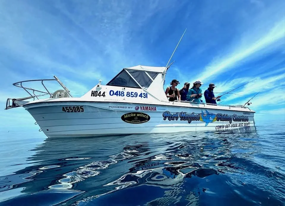 A group of people fishing from a blue and white boat on calm water under a blue sky with wispy clouds.