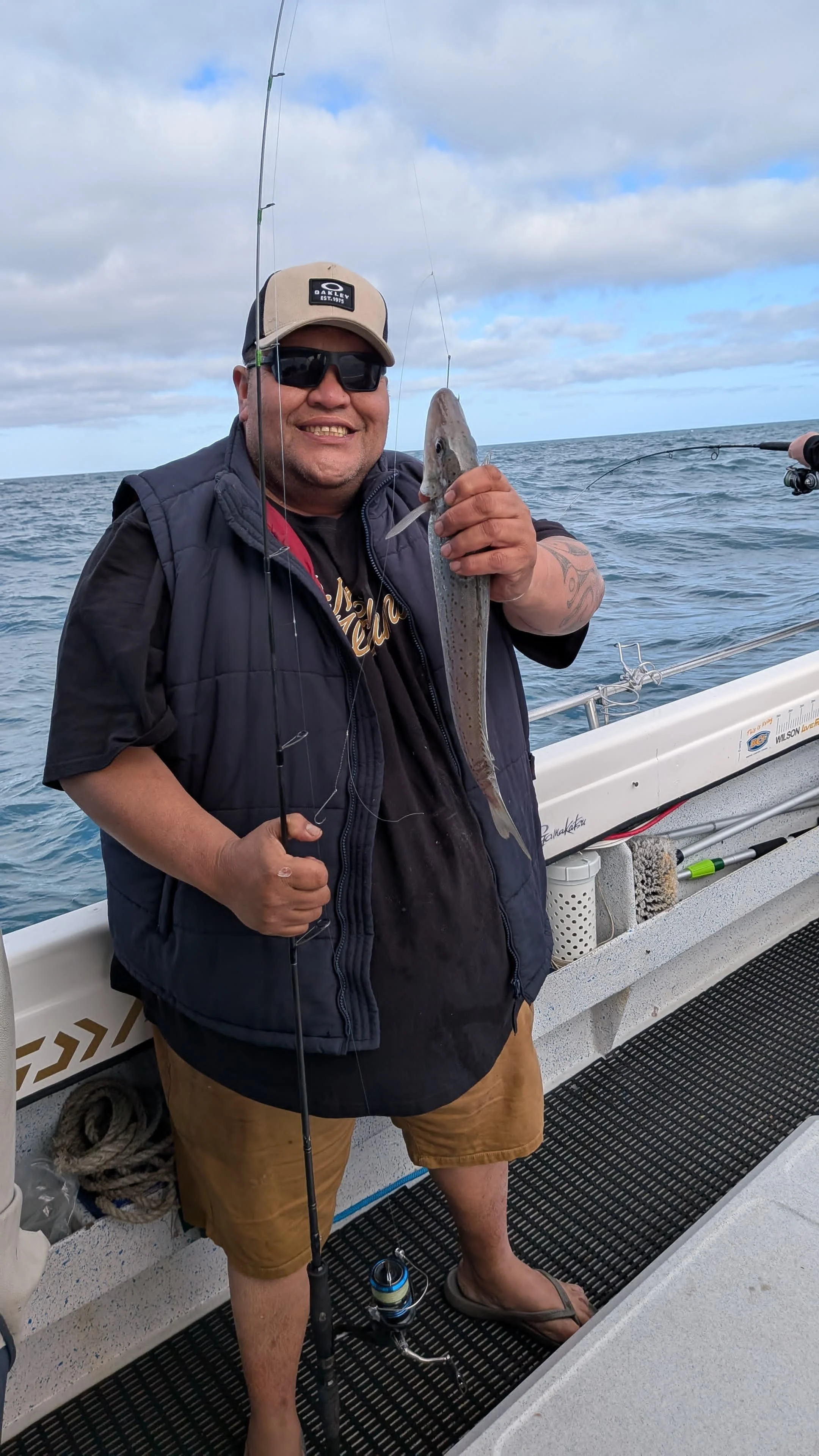 Man smiling on a boat holding a fishing rod and a small fish, with a fishing pole and ocean in the background.