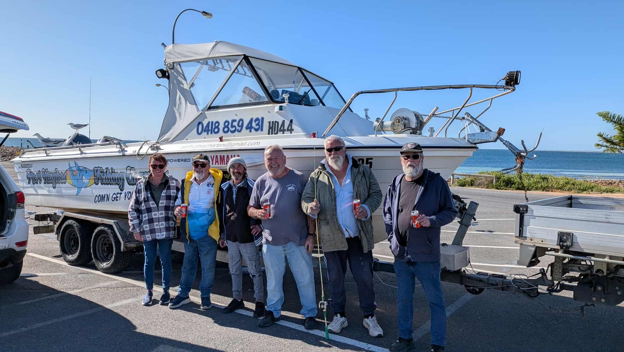Group of six people standing in front of a boat on a trailer, holding drinks, near the coast on a sunny day.