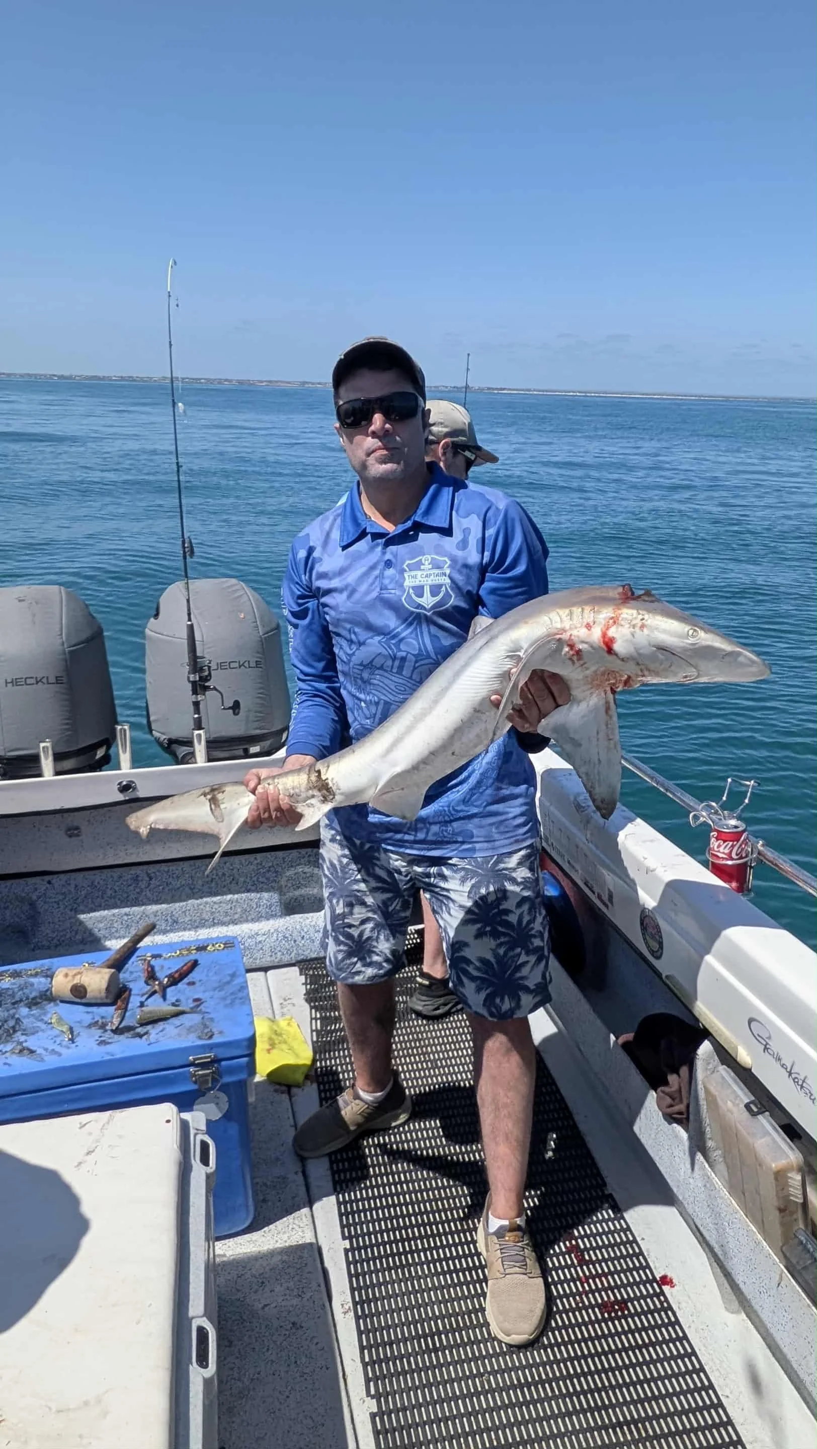 A man wearing sunglasses, a blue shirt, and patterned shorts on a boat, holding a large fish. In the background, there are fishing rods, coolers, and a calm body of water with a clear blue sky.
