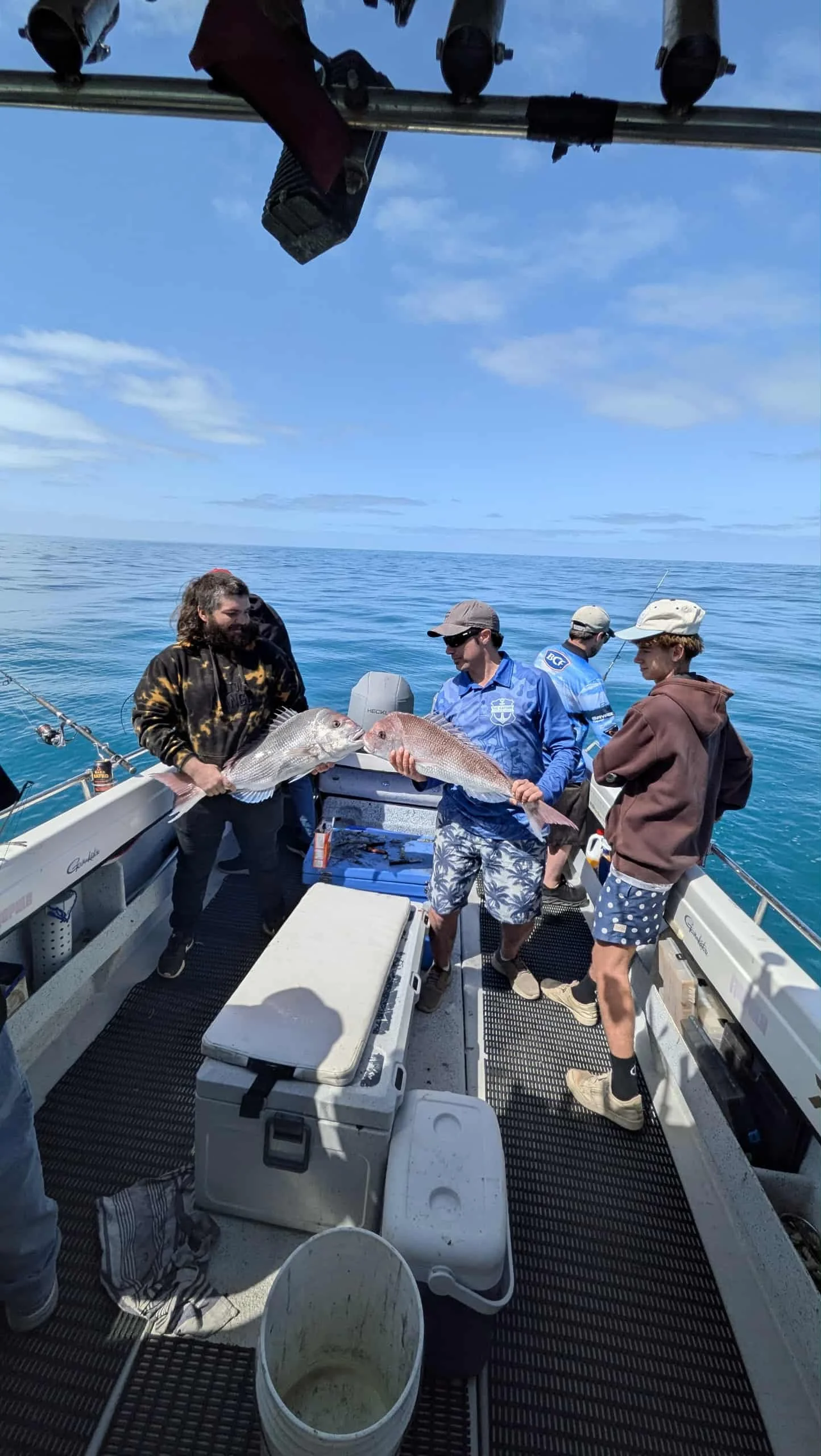 Four men on a fishing boat holding large fish, with other fishing gear visible and ocean in the background under a blue sky.