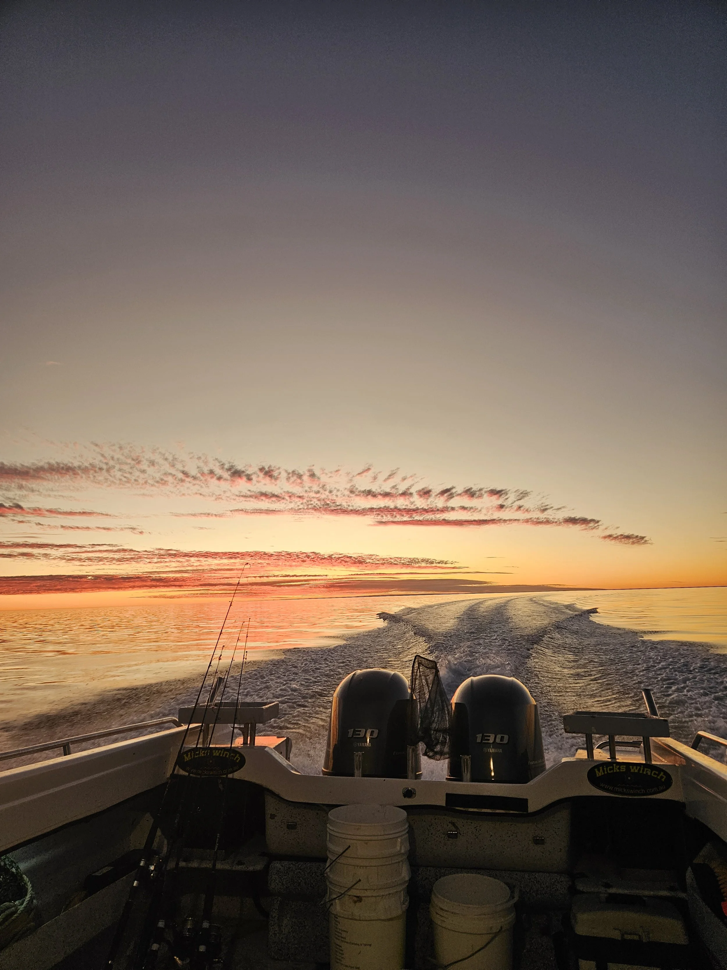 View from a boat at sunset, showing the wake in the water with the sky painted in colors of orange, pink, and purple. The boat has two outboard motors, fishing rods, and buckets visible on the deck.