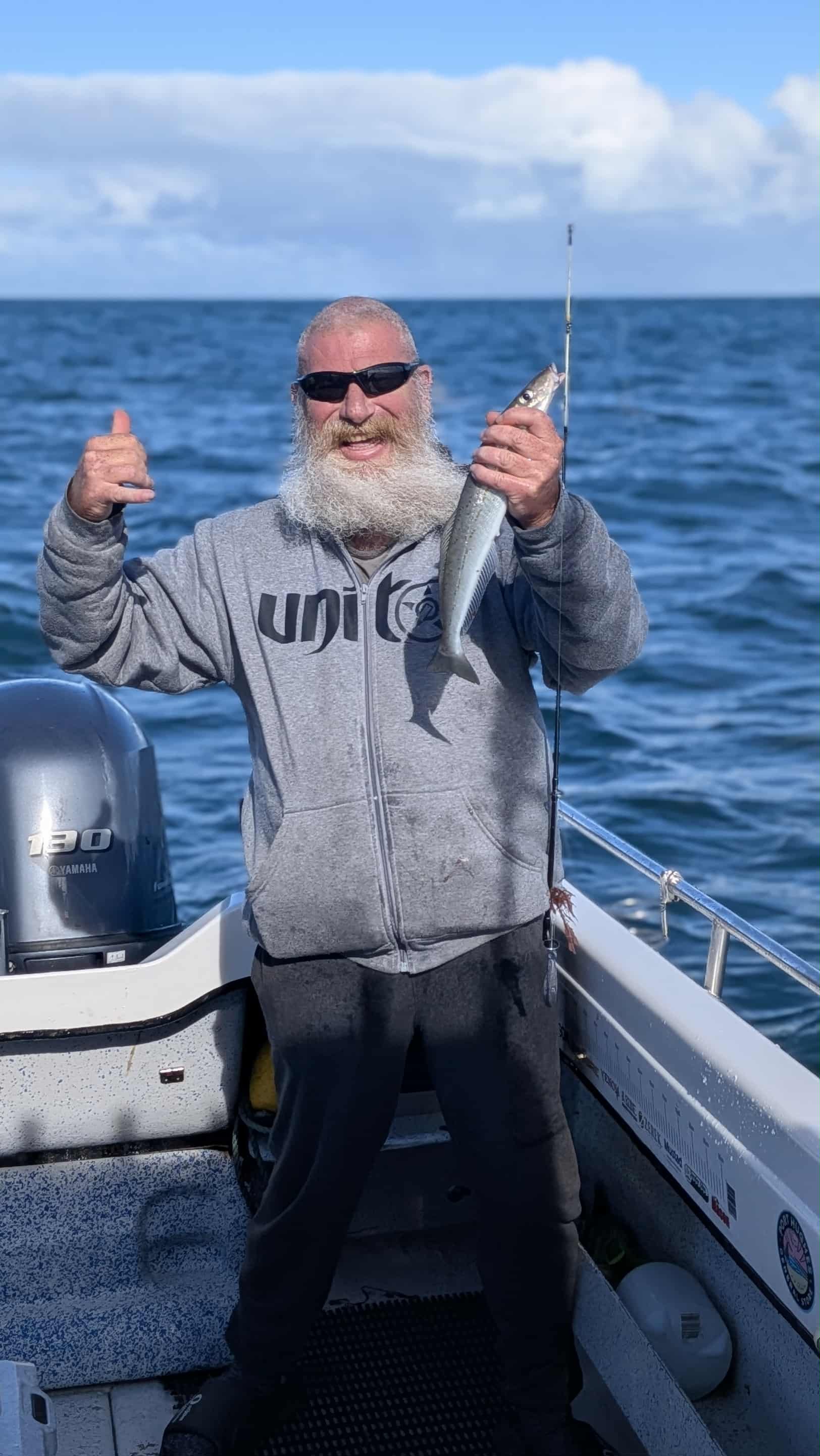 A man with a beard and sunglasses holding a fish on a boat in the ocean with a blue sky and clouds in the background.