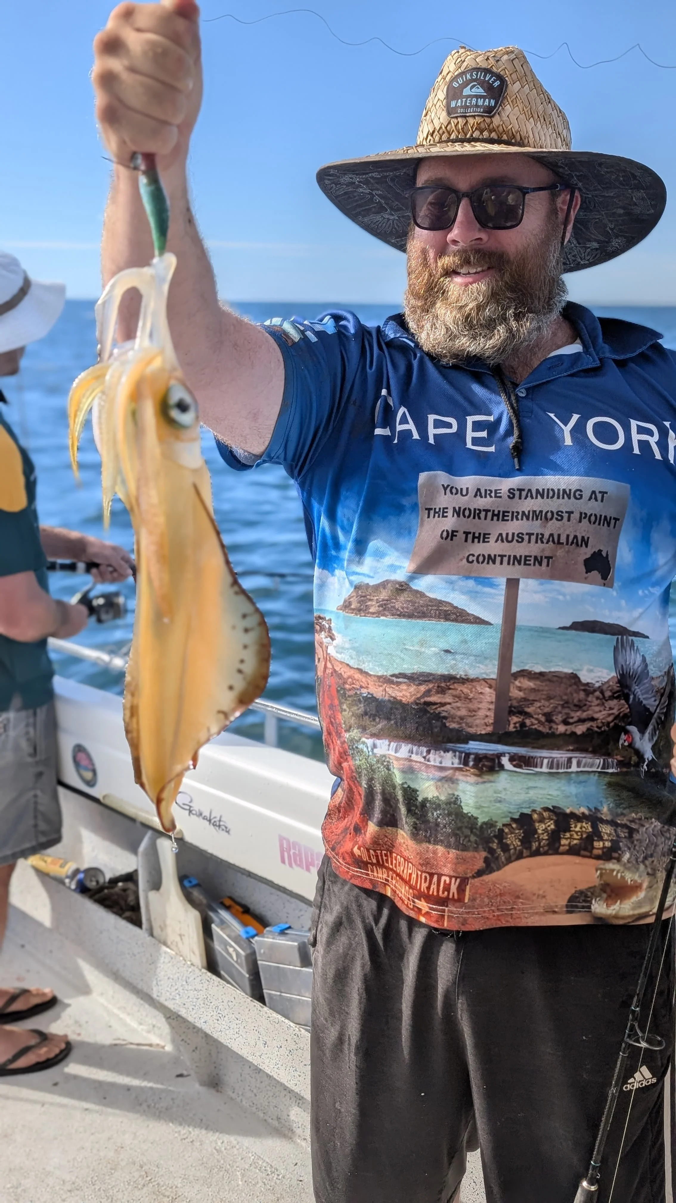 A man holding a small squid he caught while fishing on a boat. The man is wearing a wide-brimmed straw hat, sunglasses, a colorful t-shirt, and dark pants. The background shows the ocean and part of another person wearing a hat.