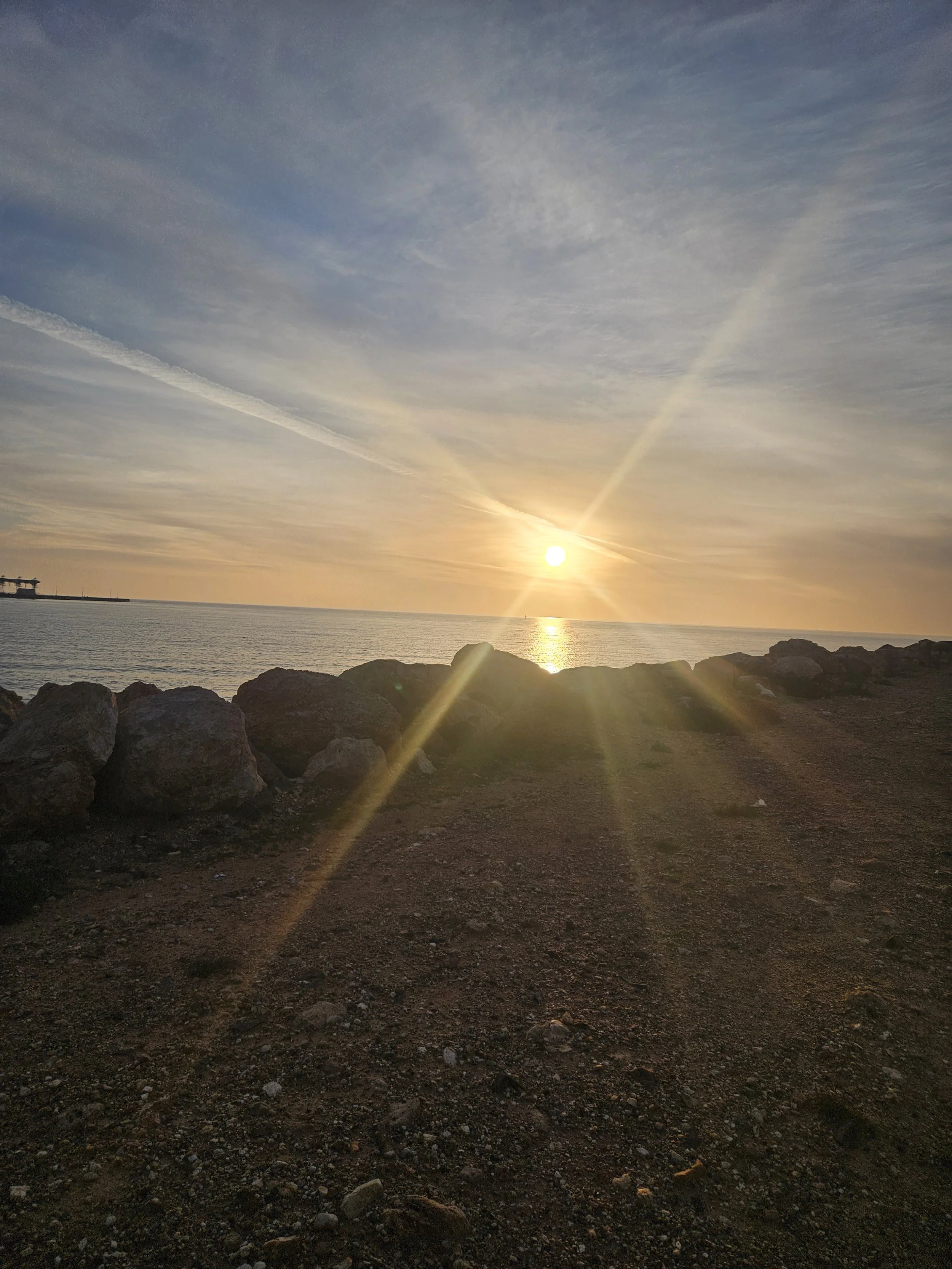 Sunset over the ocean with a rocky shoreline and a pier on the left.