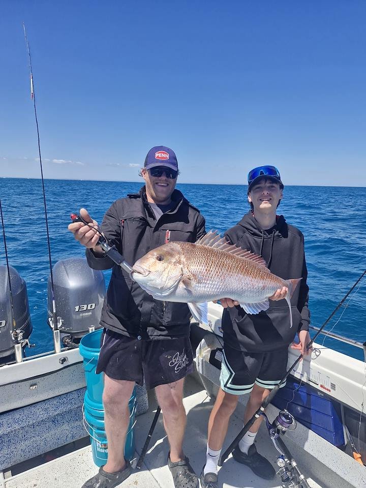 Two people on a boat holding a large fish they caught during a fishing trip, with the ocean and a clear sky in the background.
