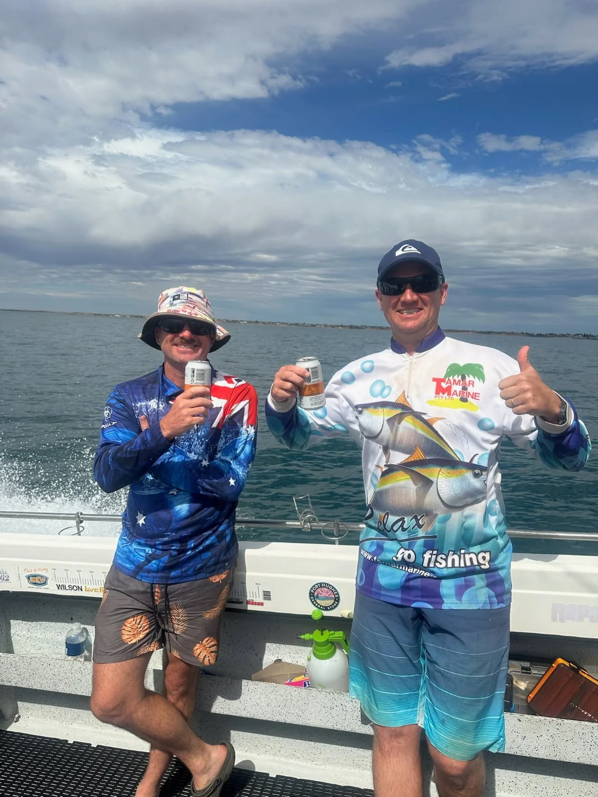 Two men on a boat holding cans of beverage, smiling and giving a thumbs-up, with a body of water and cloudy sky in the background.
