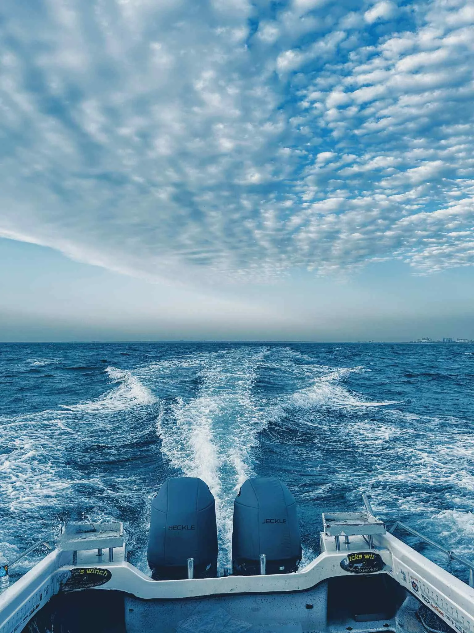 View from the back of a boat showing the ocean with waves and a partly cloudy sky. The boat's motors are visible with the words "HECKLE" and "JECKLE" on them, and there are other boat equipment visible at the bottom of the image.