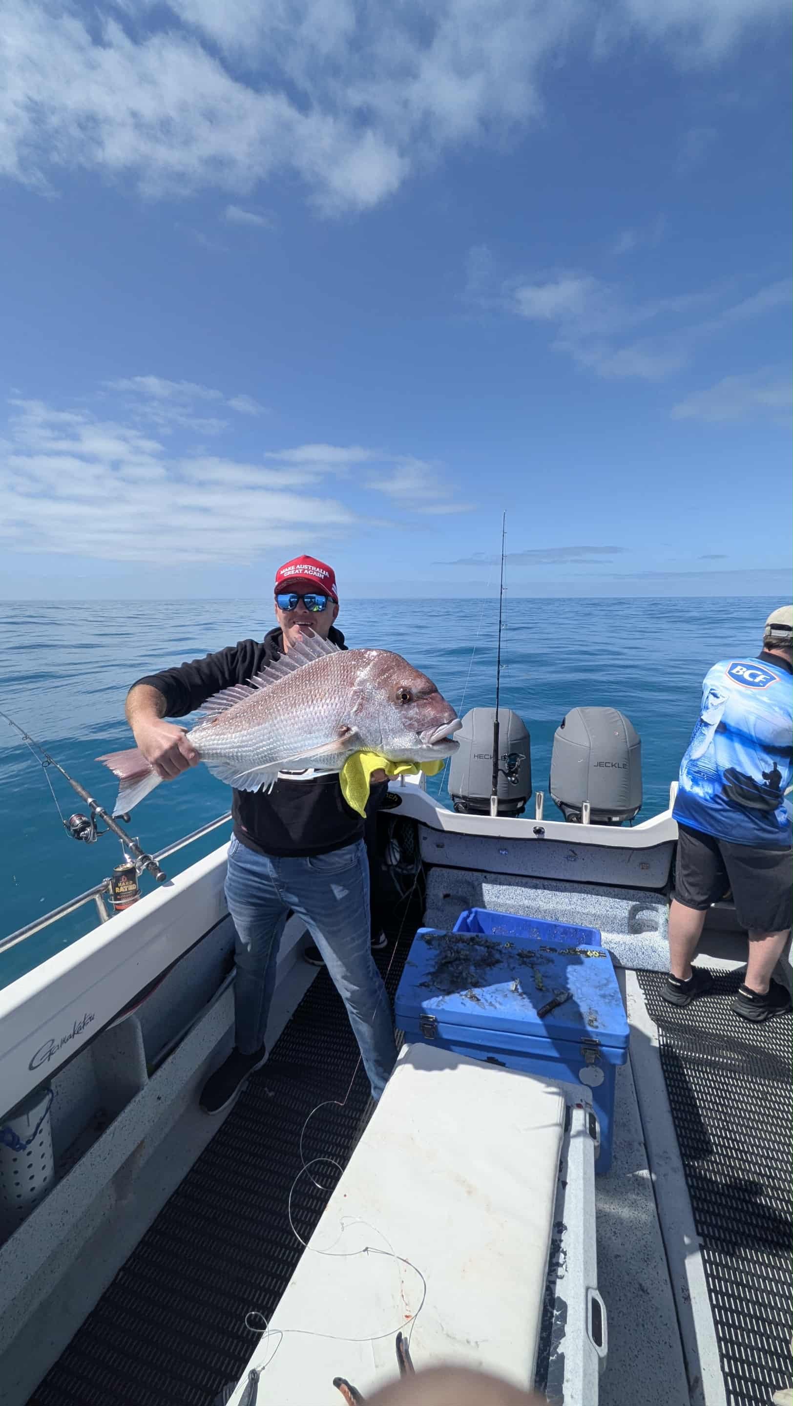 A man on a boat holding a large fish with pink and silver scales. Another person is partially visible on the right, and the boat is equipped with fishing gear and seats. The scene is on open water with a blue sky and few clouds.