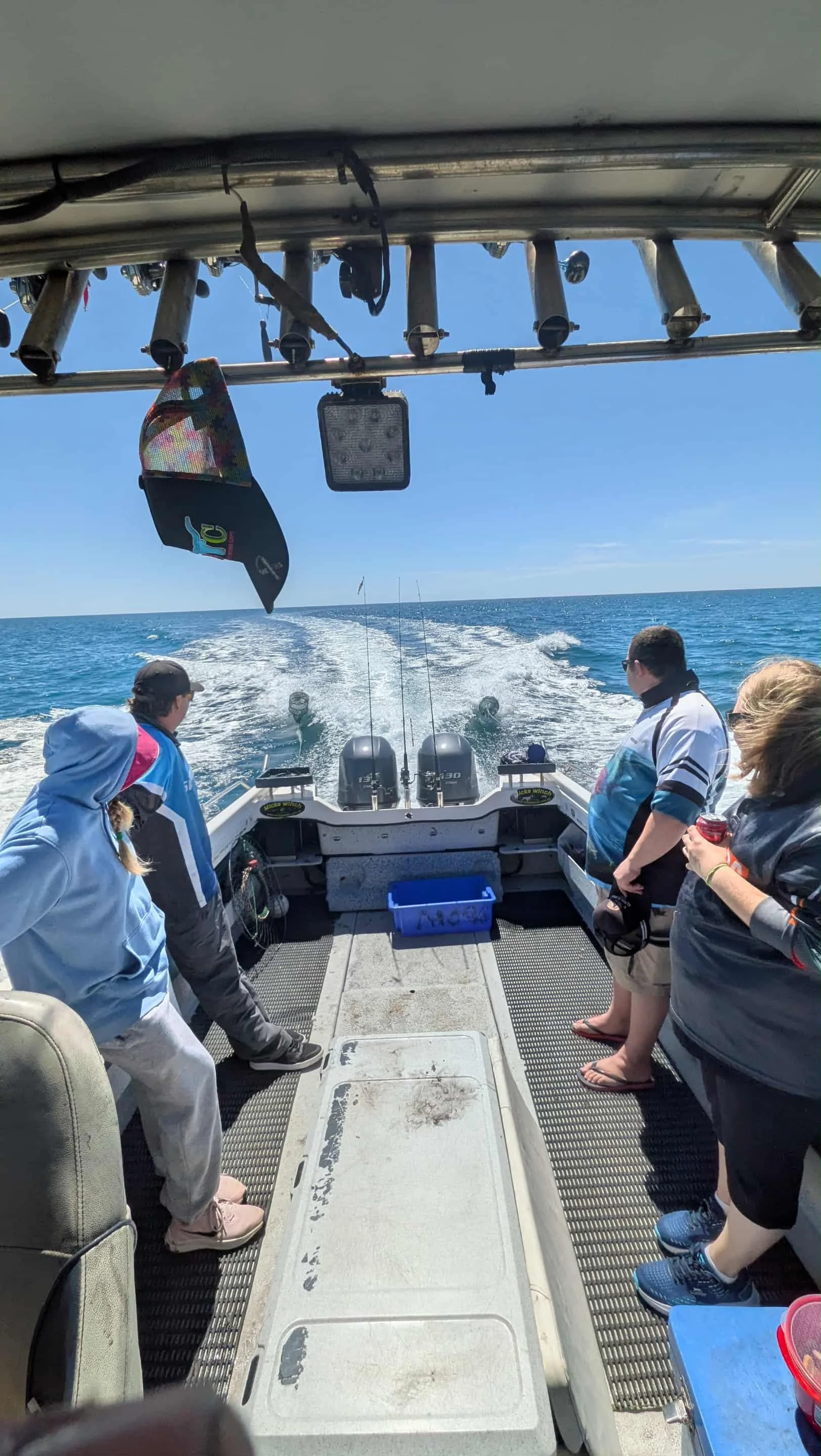 People on a boat fishing in the ocean, with fishing rods lowered into the water and a wake visible behind the boat, under a clear blue sky.