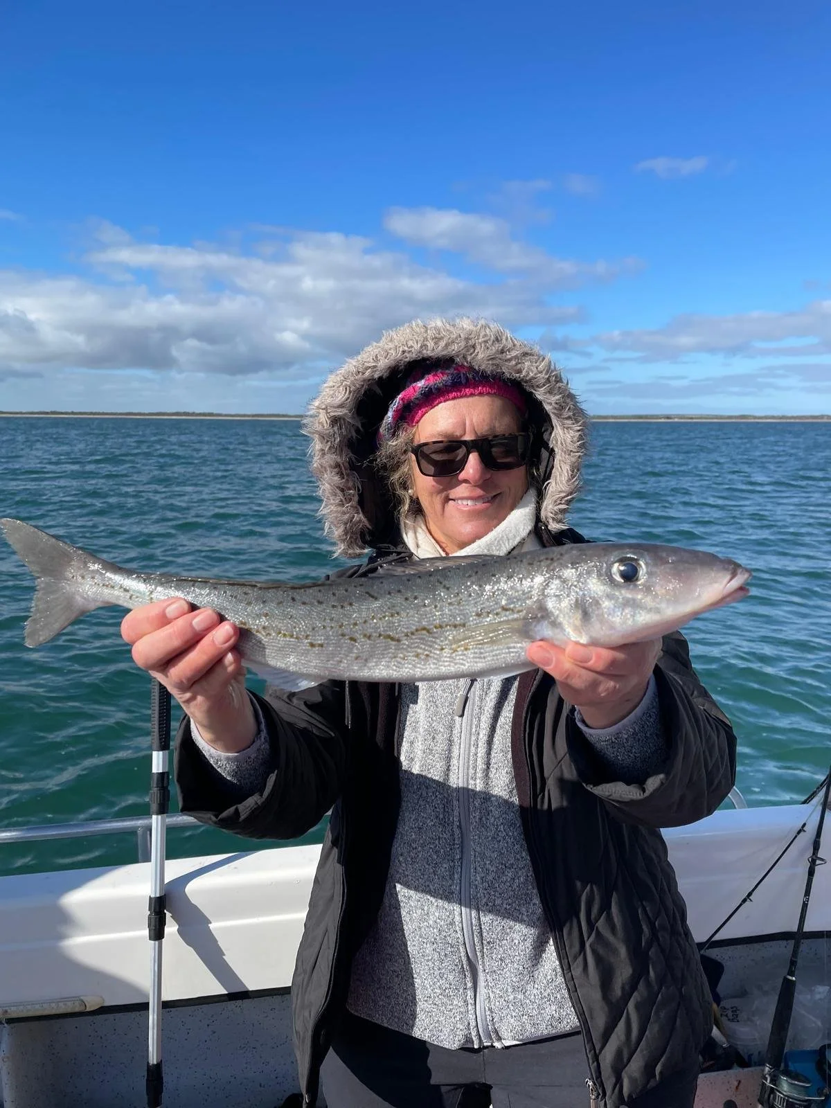 Woman in winter clothing and sunglasses holding a large fish on a boat with water and a cloudy sky in the background.