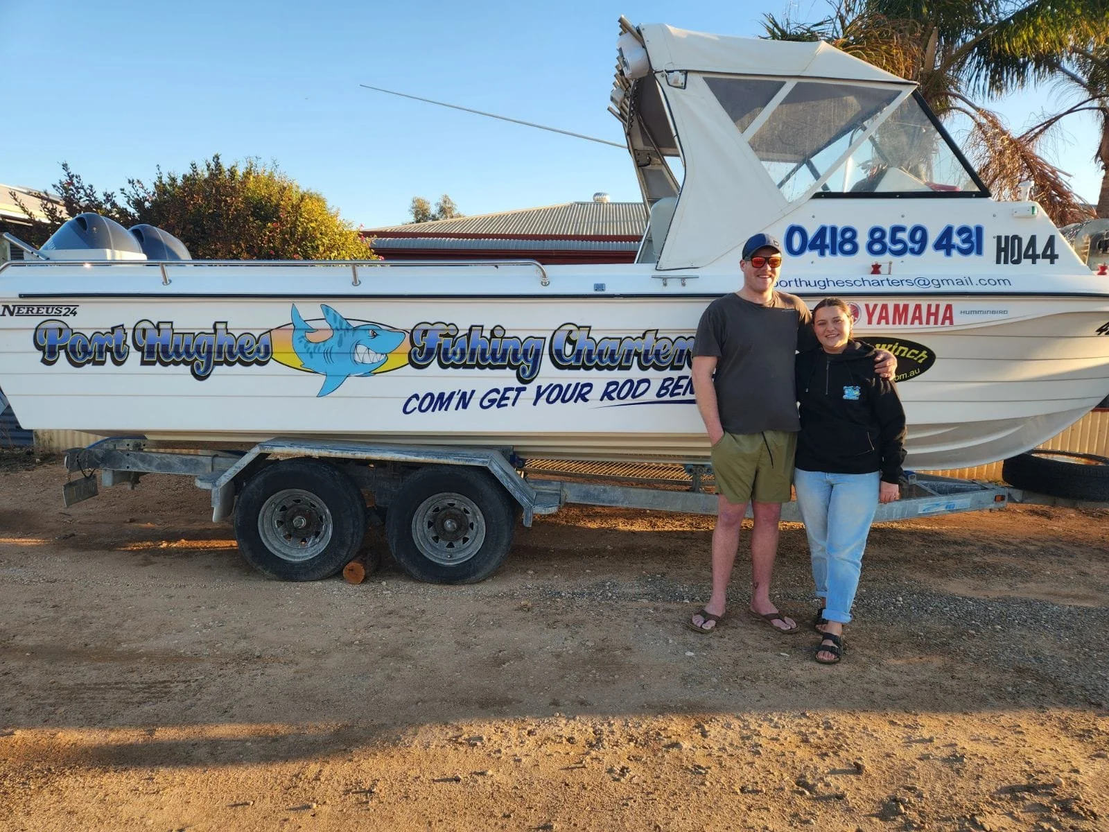 Two people standing next to a fishing boat on a trailer. The boat has a blue shark logo and text advertising Port Hughes Fishing Charters.