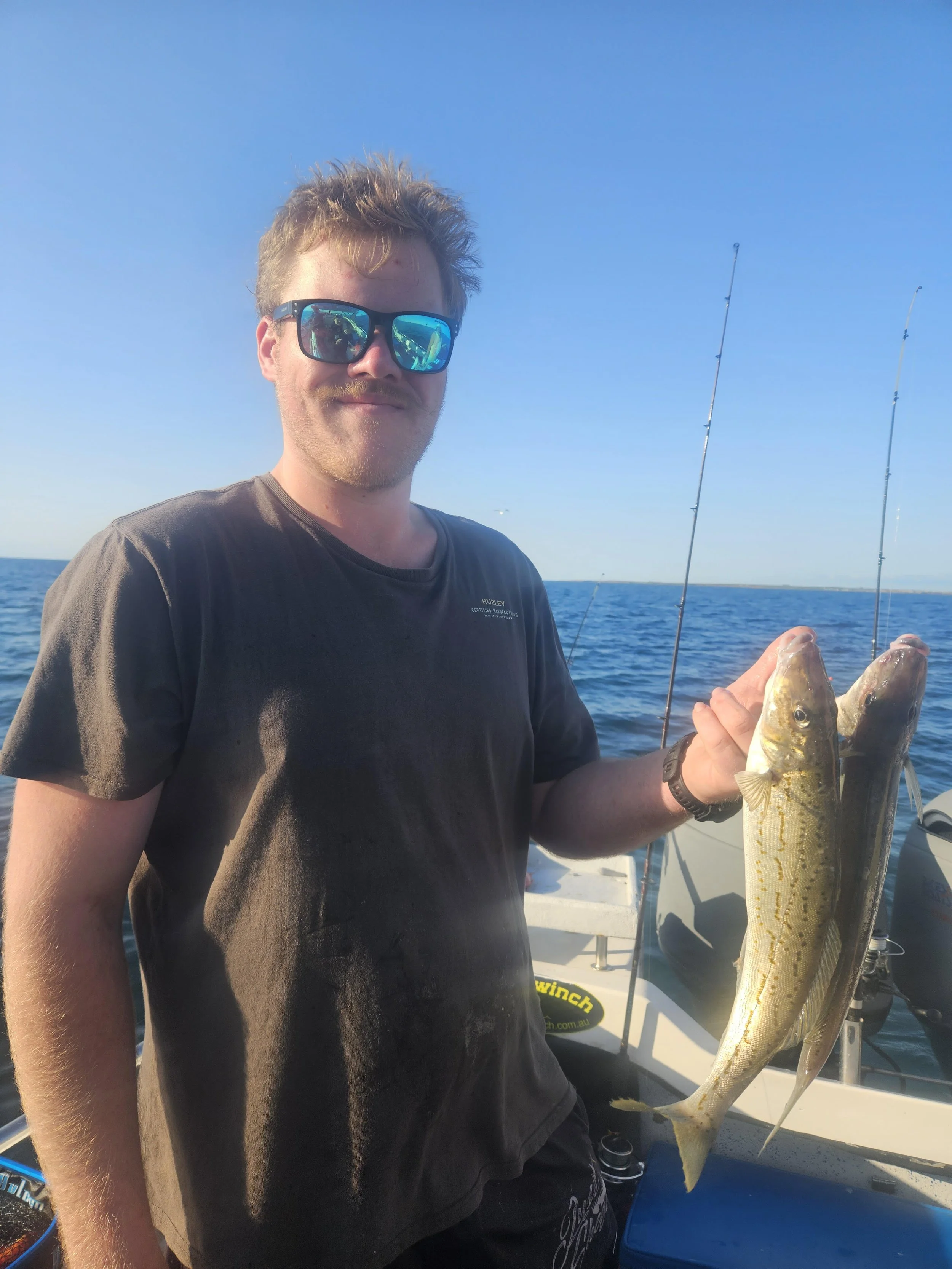 A man with sunglasses holding two fish and smiling on a boat in the open sea during daytime.