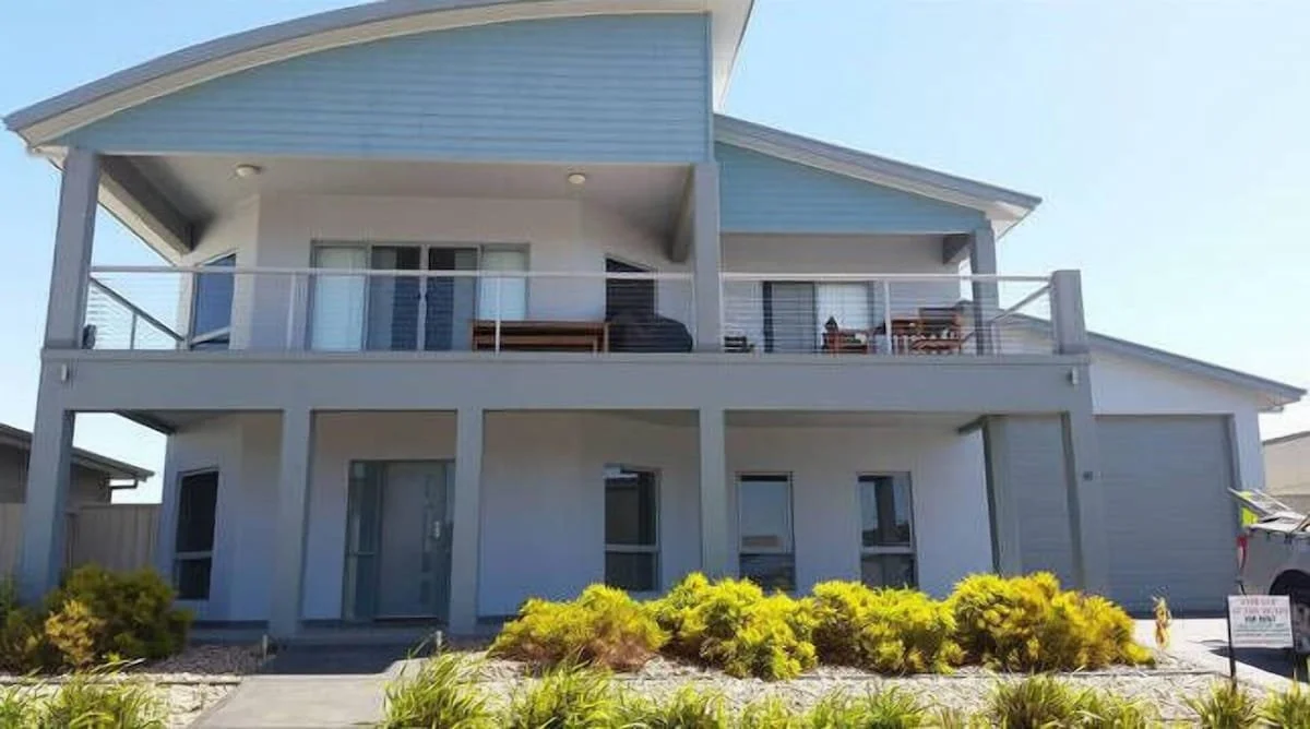 Front view of a two-story house with a light blue and white exterior, a curved roof, balcony with railing, outdoor furniture, and a landscaped yard with small bushes and plants.