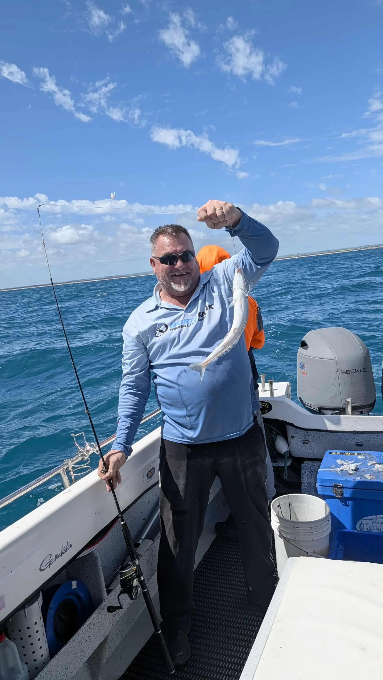 A man on a boat holding a fish he caught, dressed in outdoor fishing apparel, with a fishing rod, ocean and blue sky in the background.