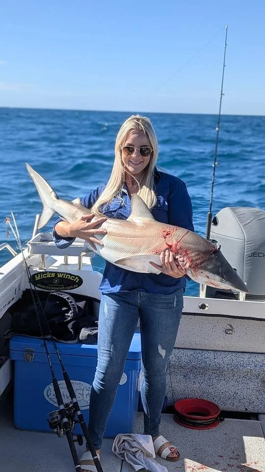 A young woman in sunglasses holding a large shark on a boat at sea, with fishing equipment and ocean in the background.