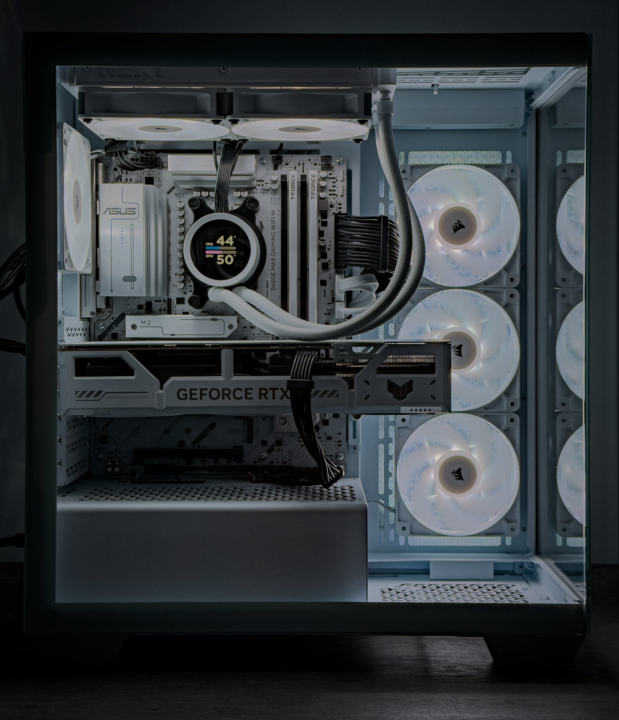 Inside a custom-built gaming PC case showing three white cooling fans, a motherboard with labeled components, a water cooling system with a circular CPU block displaying temperature readings, an NVIDIA GeForce RTX graphics card, and various black cables connecting hardware parts.